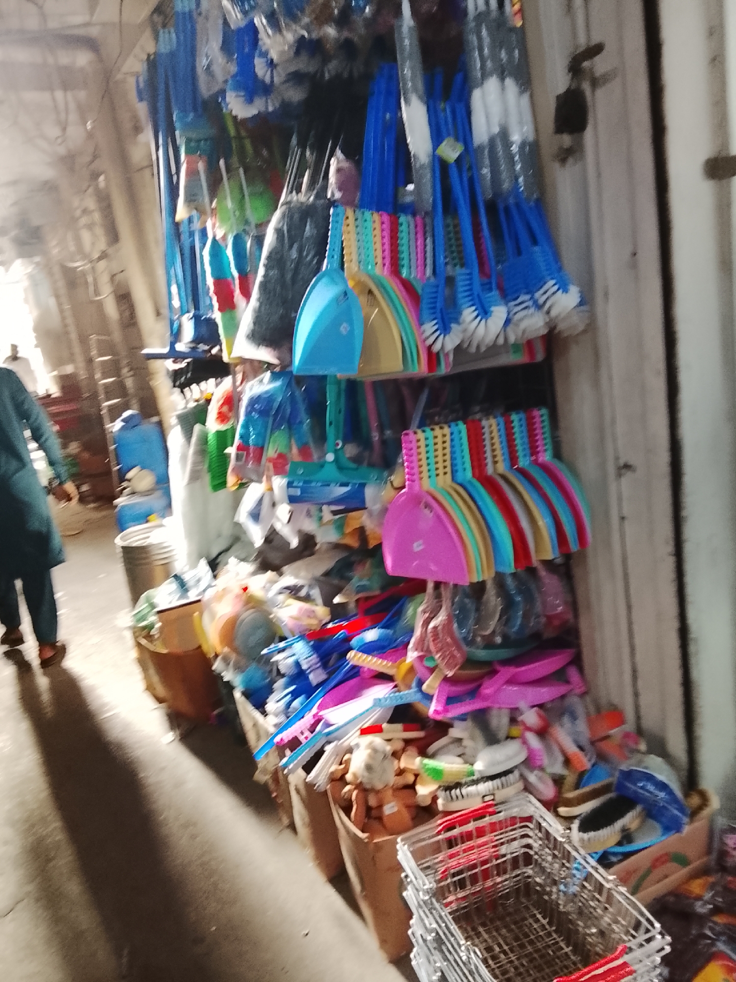 The image depicts a street market stall in Gulgasht Colony, Pakistan. The stall is densely packed with cleaning supplies, including brooms, toilet brushes, dustpans, and scrub brushes in various colors. These items hang from above and are stacked in bins. In the foreground, there are shopping baskets. In the background, a person in a teal outfit is walking away from the camera. The overall environment suggests a busy, compact, and commercially-driven street scene.