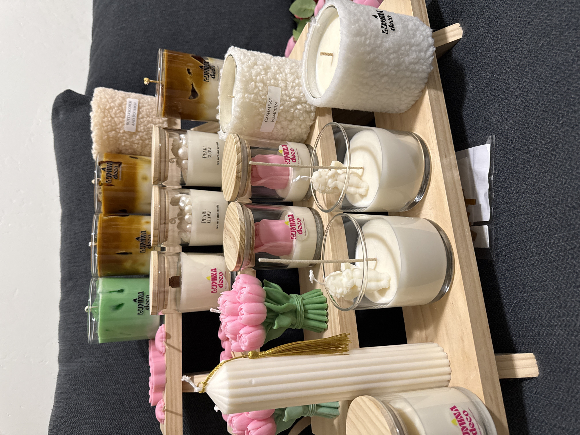 This is a top-down, eye-level, medium shot of a collection of decorative candles displayed on a light-colored wooden tiered shelf. The candles are of various shapes, sizes, and colors, with some appearing to be in glass jars and others as standalone molded candles. Notable are several cylindrical candles, some with a fluted design, a bouquet of pink tulip-shaped candles, and candles with fluffy exterior coverings. Some candles have visible wicks, and a few have labels indicating scents like "Pearl Glow" and "Cashmere Pumpkin." The background is dominated by a dark gray, textured surface, likely a couch or fabric upholstery, and a white wall. No people are visible in the scene. The lighting appears to be ambient, casting soft shadows. The image does not provide any information that would allow for its localization within Herriman, United States of America, or any other specific geographical location.