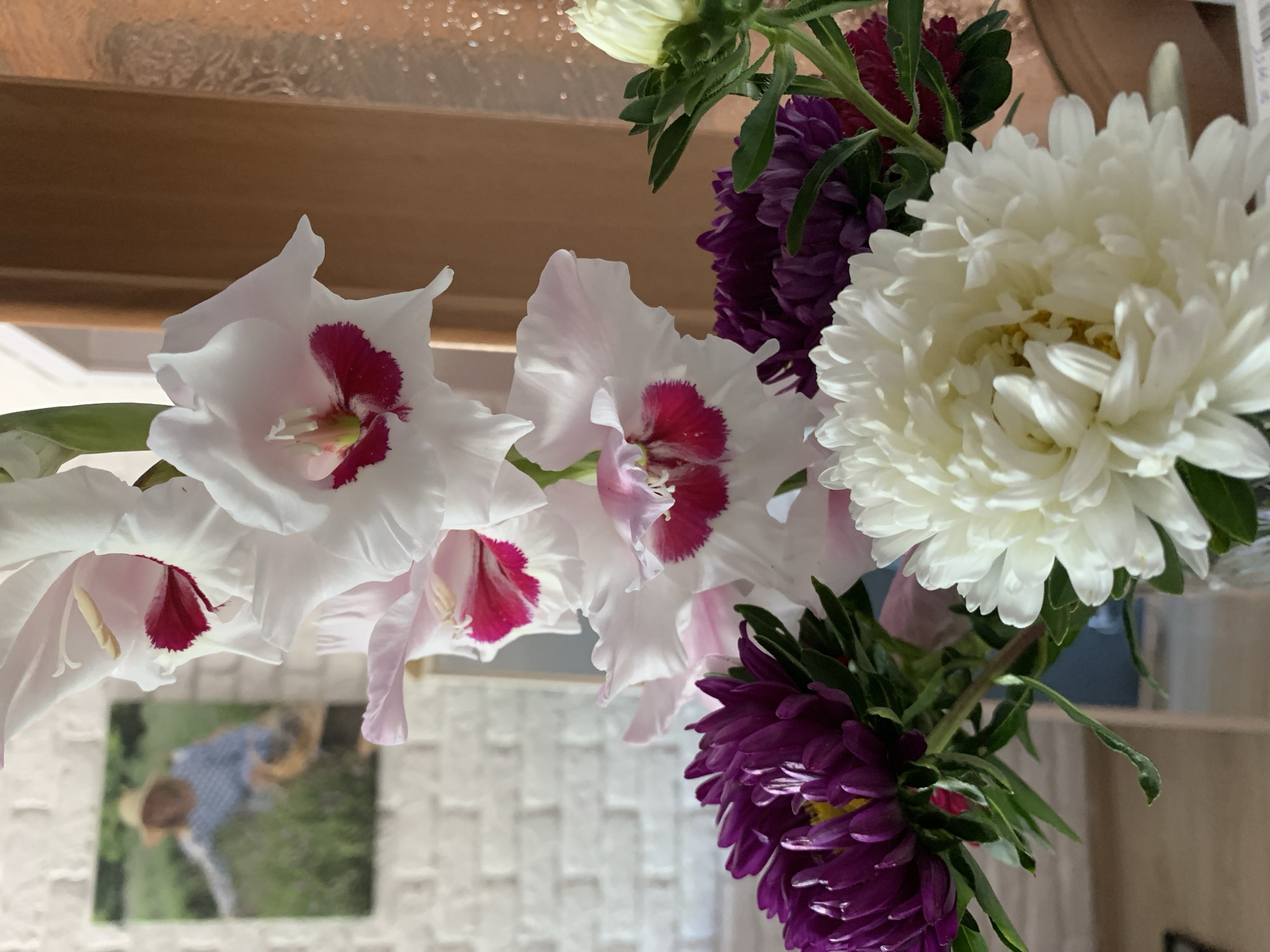 A close-up, vertical shot displays a bouquet of flowers in the foreground, with a blurred background suggesting an indoor setting. The bouquet features white gladiolus with deep pink centers, vibrant purple asters, and a large white chrysanthemum. In the background, a framed picture hangs on a white brick wall, depicting a person in a straw hat and blue shirt in a grassy field. The overall scene is brightly lit, creating a warm and inviting atmosphere. There is no indication of the city of Kapyĺ, Belarus, or any specific urban environment.