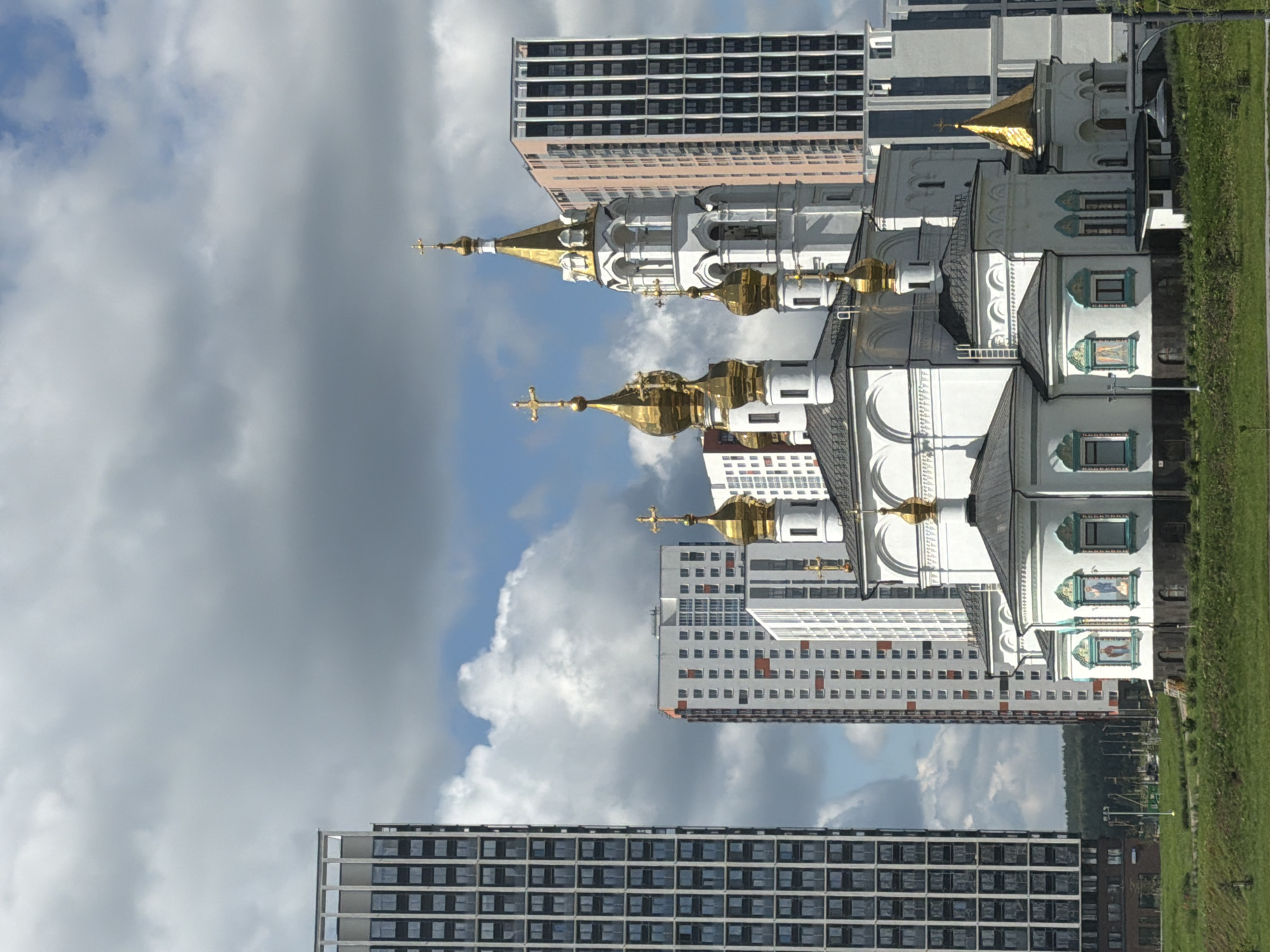 The image depicts a modern cityscape in Yekaterinburg, Russia, dominated by the contrast between a traditional Orthodox church and towering residential buildings. In the foreground, a white church with multiple golden domes and crosses stands prominently. The church features intricate architectural details, including arched windows and decorative elements. Behind the church, a series of multi-story apartment buildings rise, showcasing contemporary architectural styles with varied facades of white, red, and dark gray. The tallest building, a dark gray skyscraper with a grid-like pattern of windows, is situated on the left side of the image. The sky is partially cloudy, with patches of blue visible. A strip of green grass and vegetation forms the immediate foreground, suggesting an urban park or green space. The overall scene conveys a juxtaposition of historical and modern urban development within Yekaterinburg.