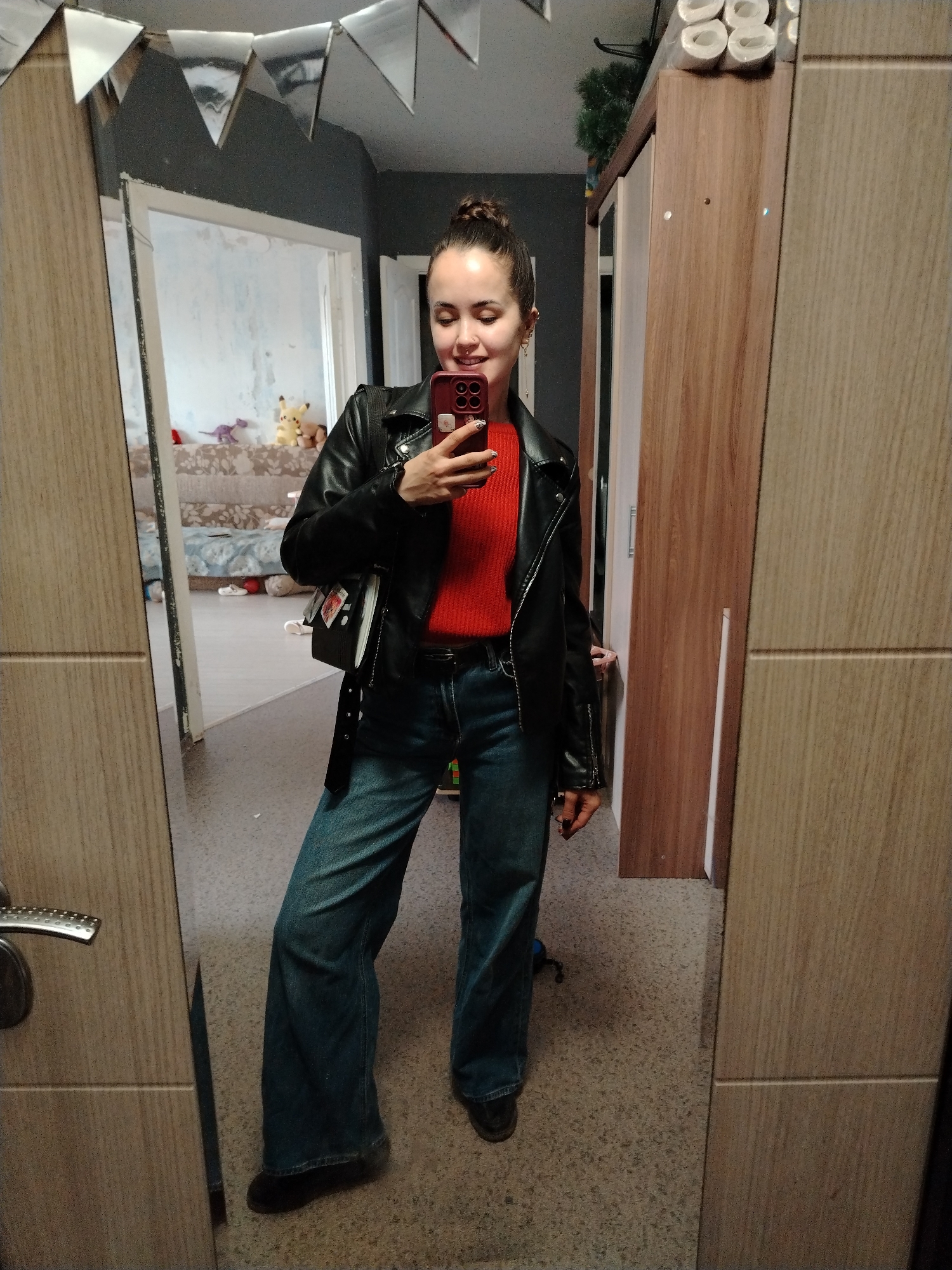 A young woman is taking a selfie in a mirror in what appears to be a residential space. She has her hair in a bun and is wearing a red sweater, a black leather jacket, and blue jeans. She is holding a phone with a red case. The background includes a room with toys, and a brown wardrobe, toilet paper, and greenery are visible in the corner. Silver triangular decorations hang from the ceiling. The location appears to be indoors in a room that is part of an apartment or house. The city, Noviy, Russia, is not apparent from the image.