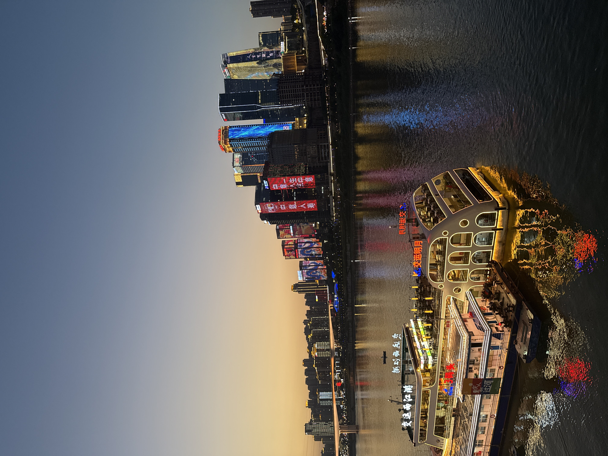 The image depicts a twilight scene in Yuzhong District, China, featuring a wide riverfront lined with illuminated skyscrapers. In the foreground, two large, brightly lit tour boats are docked. The boat on the left has multiple decks with windows, and the text "安运两江游" (An Yun Liang Jiang You) is visible on its side. The boat on the right has a distinctive curved, multi-story structure with arched windows, and the text "长运明月" (Chang Yun Ming Yue) is displayed. A smaller barge is tied to the front of the leftmost boat. The river reflects the vibrant lights of the boats and the distant cityscape. Across the river, a dense collection of modern skyscrapers dominates the skyline, many of which are adorned with colorful LED lights and illuminated advertisements. A long bridge spans the river in the middle ground, further contributing to the urban landscape. The sky transitions from a warm, orangey glow near the horizon to a deep blue overhead, indicating the transition from sunset to evening. The overall impression is one of a bustling and dynamic urban environment at night.