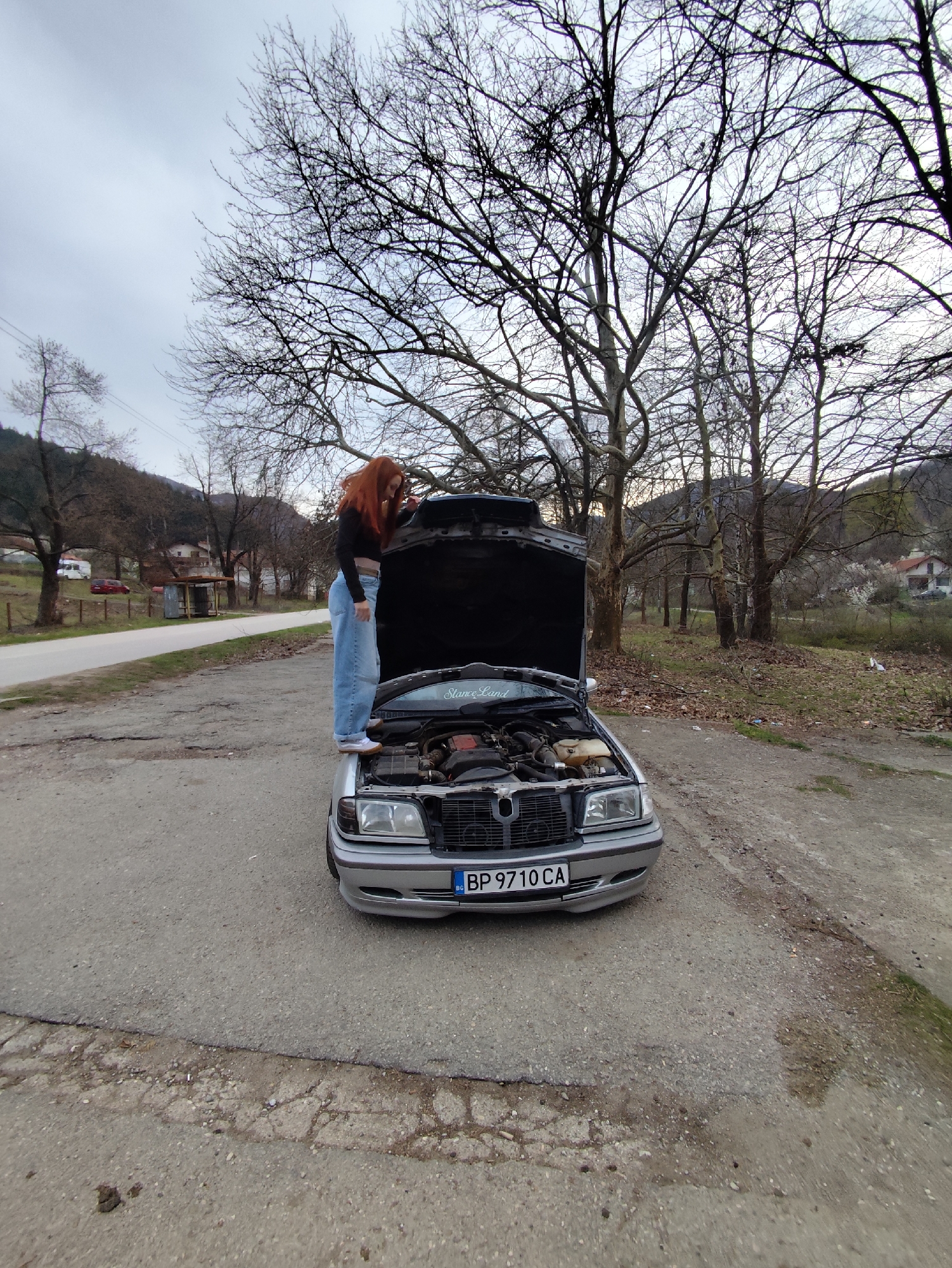 A silver Mercedes-Benz is parked on a paved surface in an outdoor location with an open hood. A young woman with long red hair stands on the car's hood, leaning over to look at the engine. She is wearing a black top and light blue jeans. The car's license plate reads "BP 9710 CA". In the background, there is a road, trees without leaves, and buildings. The scene is in Zgorigrad, Bulgaria.