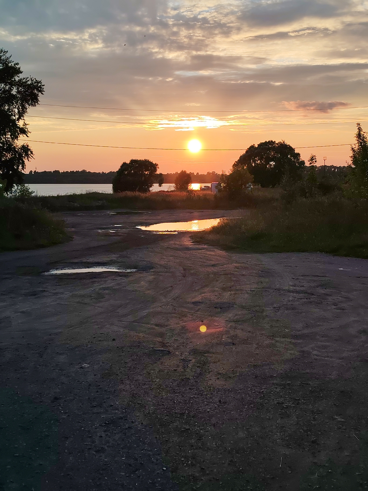 The image depicts an outdoor scene at sunset, featuring a body of water, trees, and a dirt road. The sun is visible on the horizon, casting a warm glow and reflecting off puddles on the road. Power lines are present across the sky. The environment appears rural or undeveloped, with no visible people or signs of urban activity.