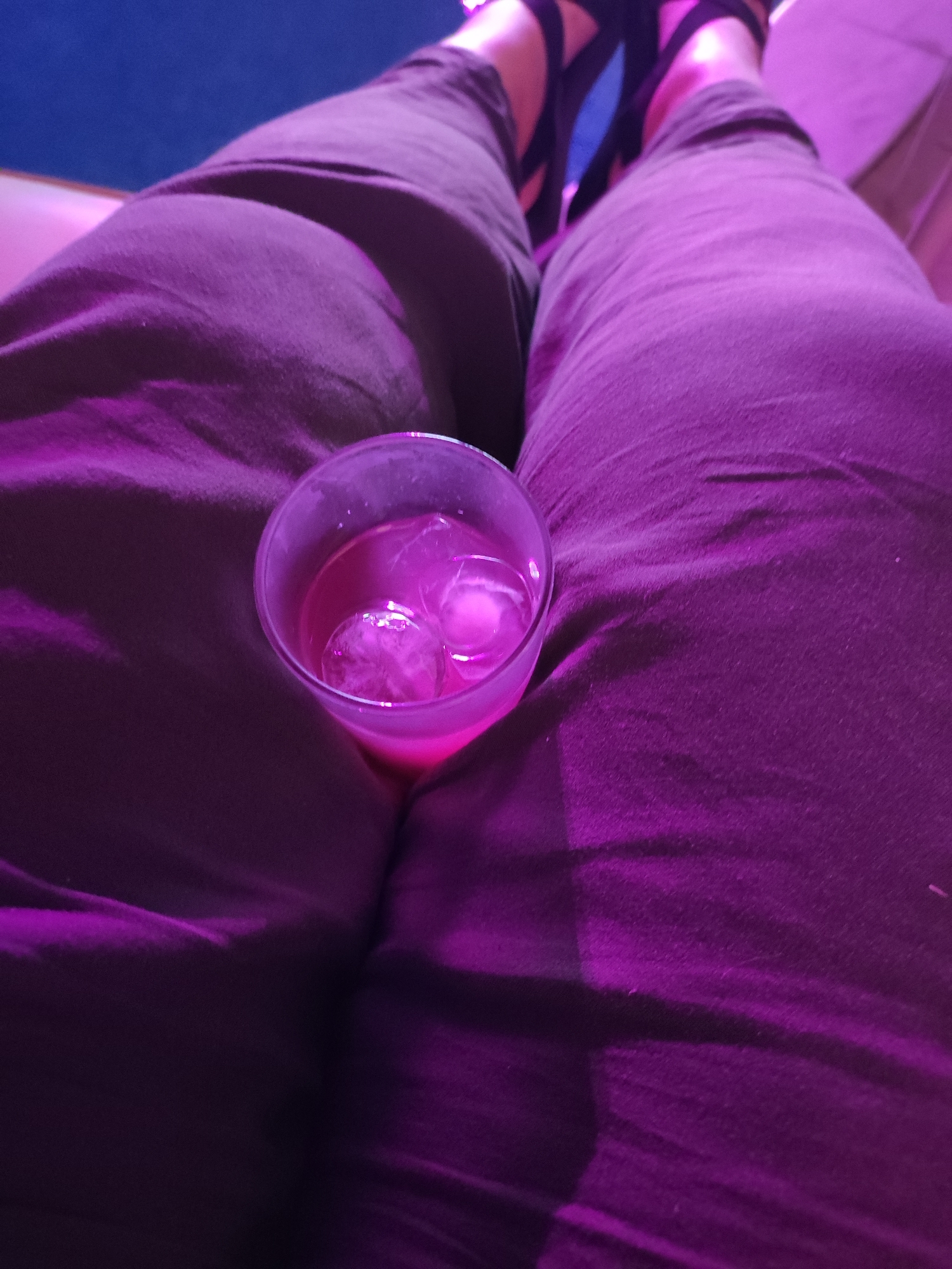 The image shows a close-up perspective of a person's lap, draped with dark purple fabric. A clear glass filled with a pink beverage and ice cubes rests on the fabric. The scene is illuminated by purple lighting, casting shadows and highlighting the textures of the clothing. The background is out of focus, appearing as a deep blue. The setting is likely indoors, possibly a bar or lounge in the Municipal Unit of Rhodes, Greece, given the context provided. No specific individuals are clearly identifiable, but the image implies the presence of at least one person.