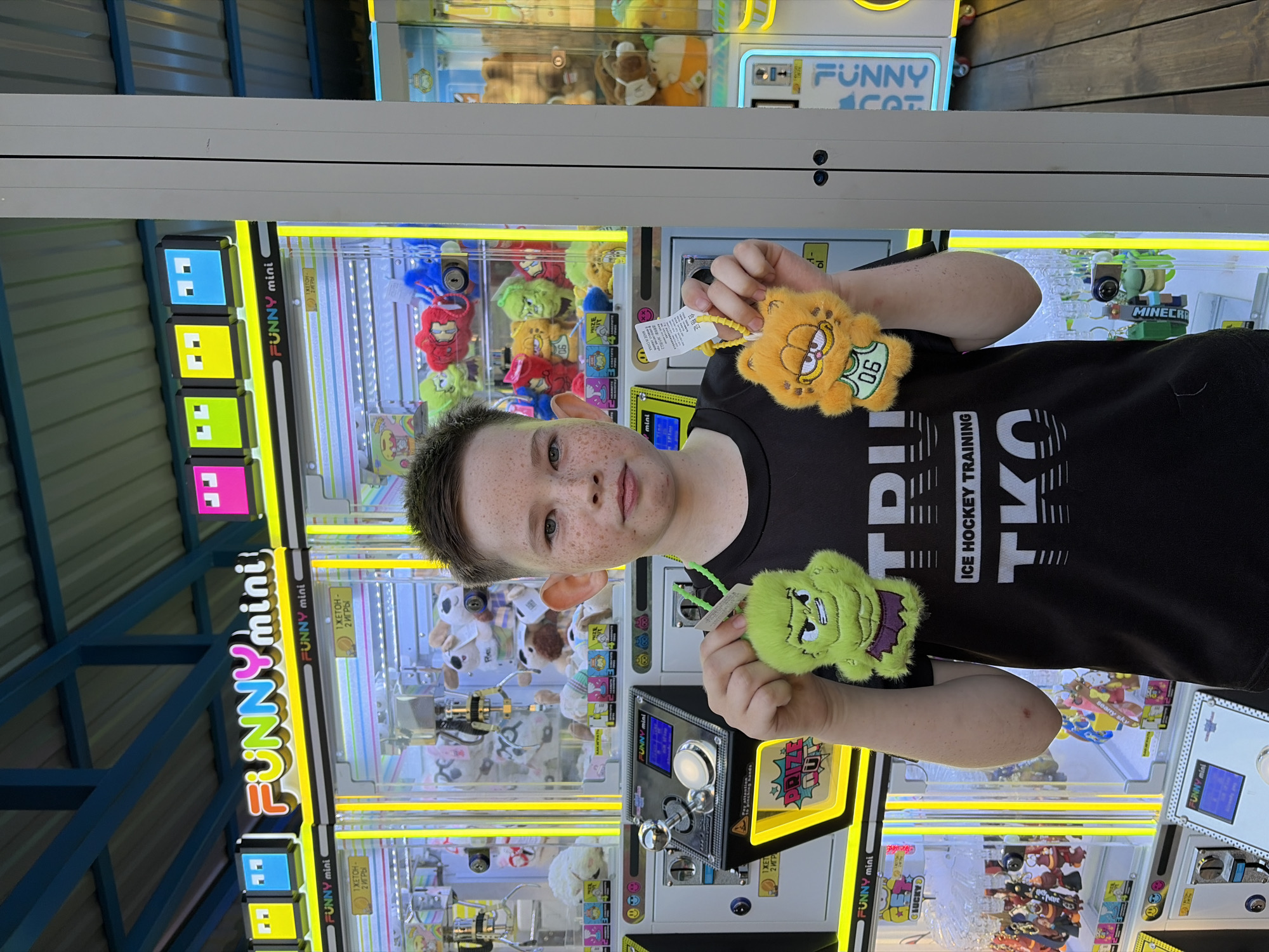 A young boy with freckles, wearing a black t-shirt, stands in front of a row of claw machines. He holds up two plush keychain toys, one green and monstrous, the other orange and resembling Garfield. The claw machines are brightly lit with yellow and blue accents, featuring the "FUNNY mini" logo and various small plush toys inside. The background shows a covered outdoor area with a metal roof structure and wooden decking. This scene is located at an amusement area or arcade in Chaikovsky, Russia.