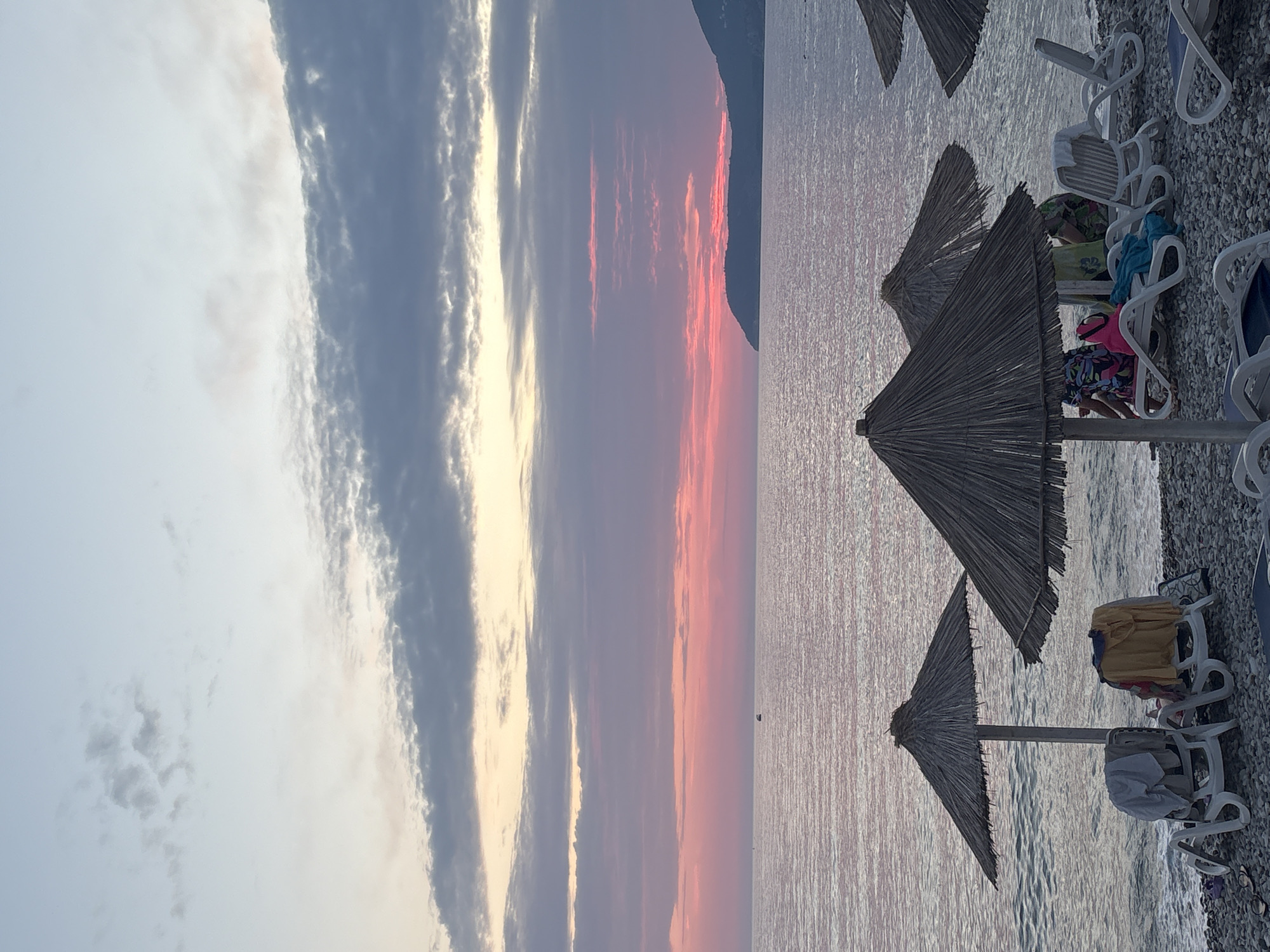 This is a media file of a beach scene in Bar, Montenegro. The image features a calm sea under a sky filled with colorful clouds at sunset. The sun is low on the horizon, casting a warm glow across the water and sky, with hues of pink, orange, and purple visible.
On the shore, there are several white lounge chairs and thatched umbrellas. Some of the chairs are occupied by people, and various beach items such as towels and bags are scattered around them. The beach itself is covered in pebbles.
The scene suggests a relaxing atmosphere, likely at the end of a summer day. The calm water and the soft lighting of the sunset contribute to a peaceful and serene mood. The presence of lounge chairs and umbrellas indicates that this is a developed beach area, possibly a resort or public beach.
No visible text is present in the image. The orientation of the image is vertical.