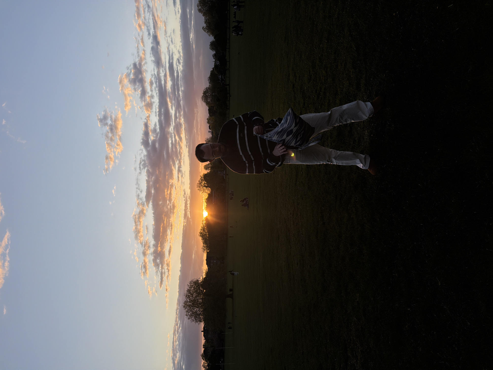 A man stands on a grassy field in London, United Kingdom, silhouetted against a sunset. He wears a striped sweater and light-colored pants, holding a patterned bag. The sky is a mix of blue and orange hues, with clouds illuminated by the setting sun. People are scattered across the field in the background. Trees and buildings are visible along the horizon.