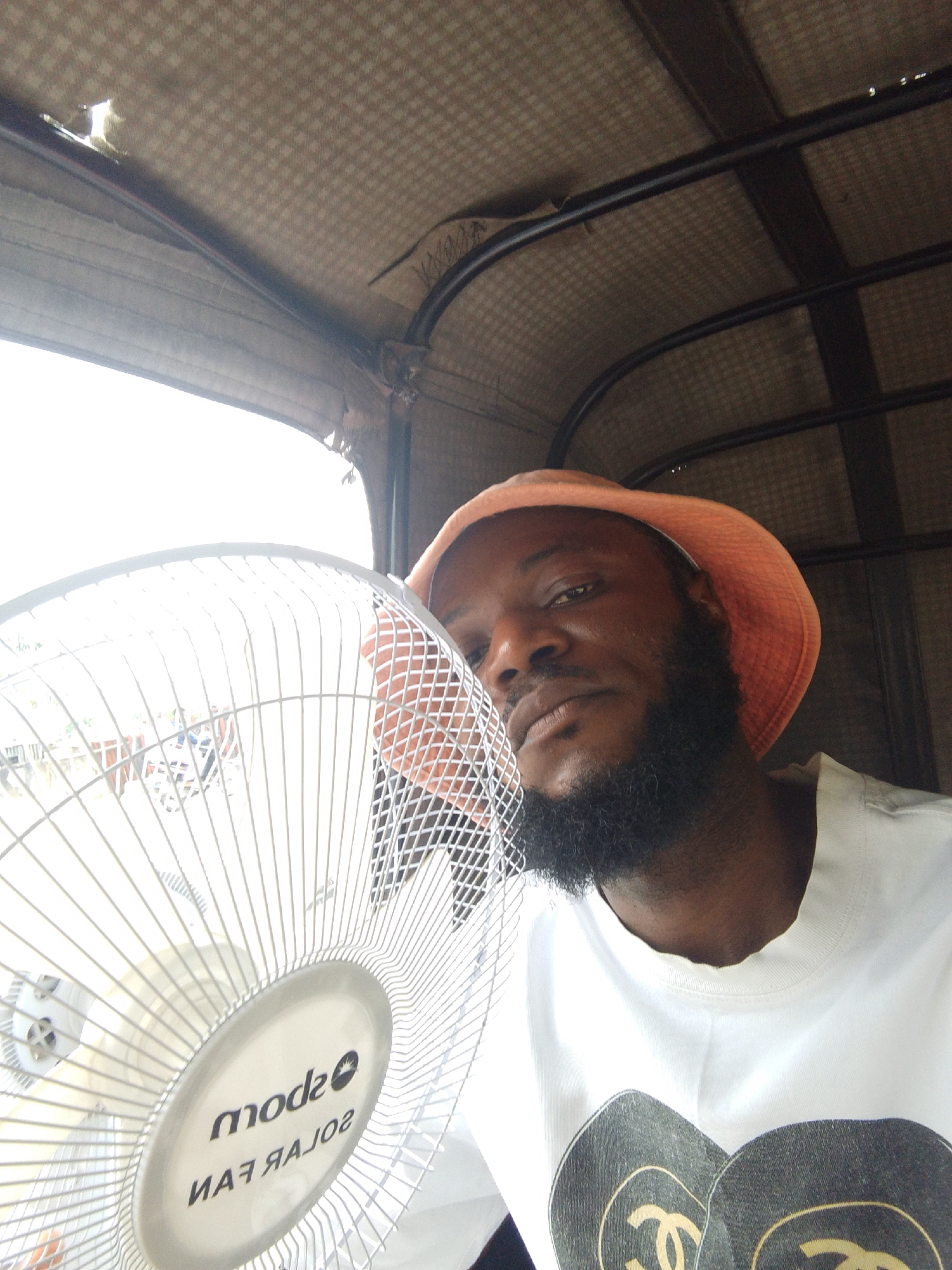 A man is inside a vehicle, possibly an auto-rickshaw, in Lokoja, Nigeria. He is holding a white electric fan close to his face. The vehicle's interior shows a worn, patterned ceiling. The lighting suggests it's daytime. The man wears a peach-colored bucket hat and a white T-shirt with a logo. He has a dark beard. The fan is a "mode solar fan." A window is visible in the background.