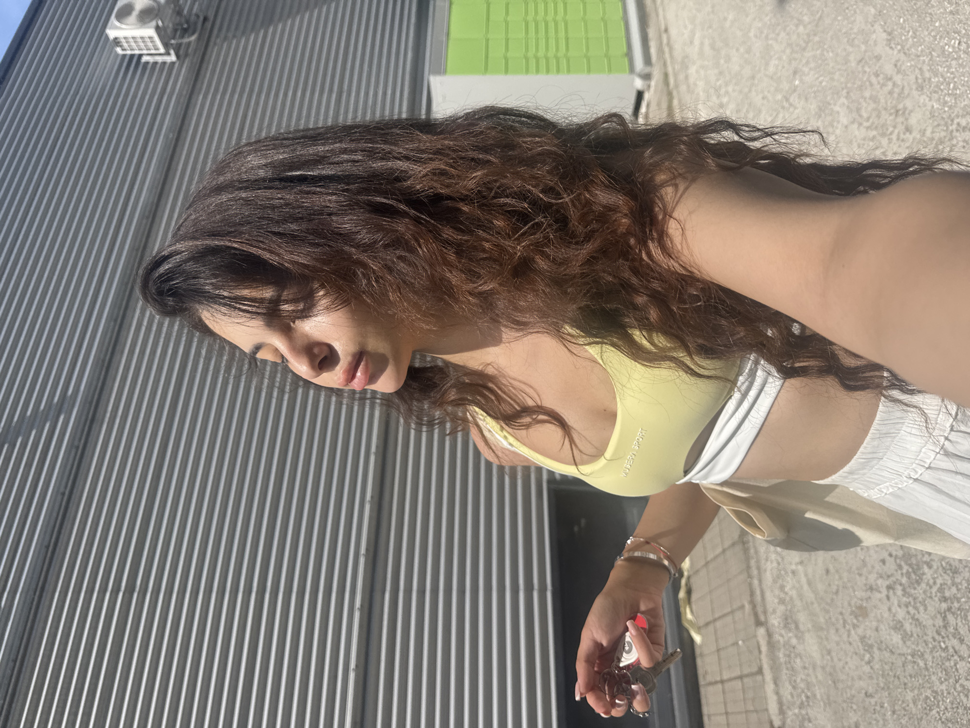 A young woman with long, dark, wavy hair stands in front of a corrugated metal wall. She is wearing a yellow and white sports bra and white pleated shorts. Her eyes are closed, and her face is tilted upwards, likely towards the sun. In her right hand, she holds a set of keys. To her left, a cream-colored bag is visible. The background consists of the metal wall, an outdoor air conditioning unit, and a green and white structure. The scene appears to be outdoors, possibly in an urban area. The lighting suggests it is daytime.
