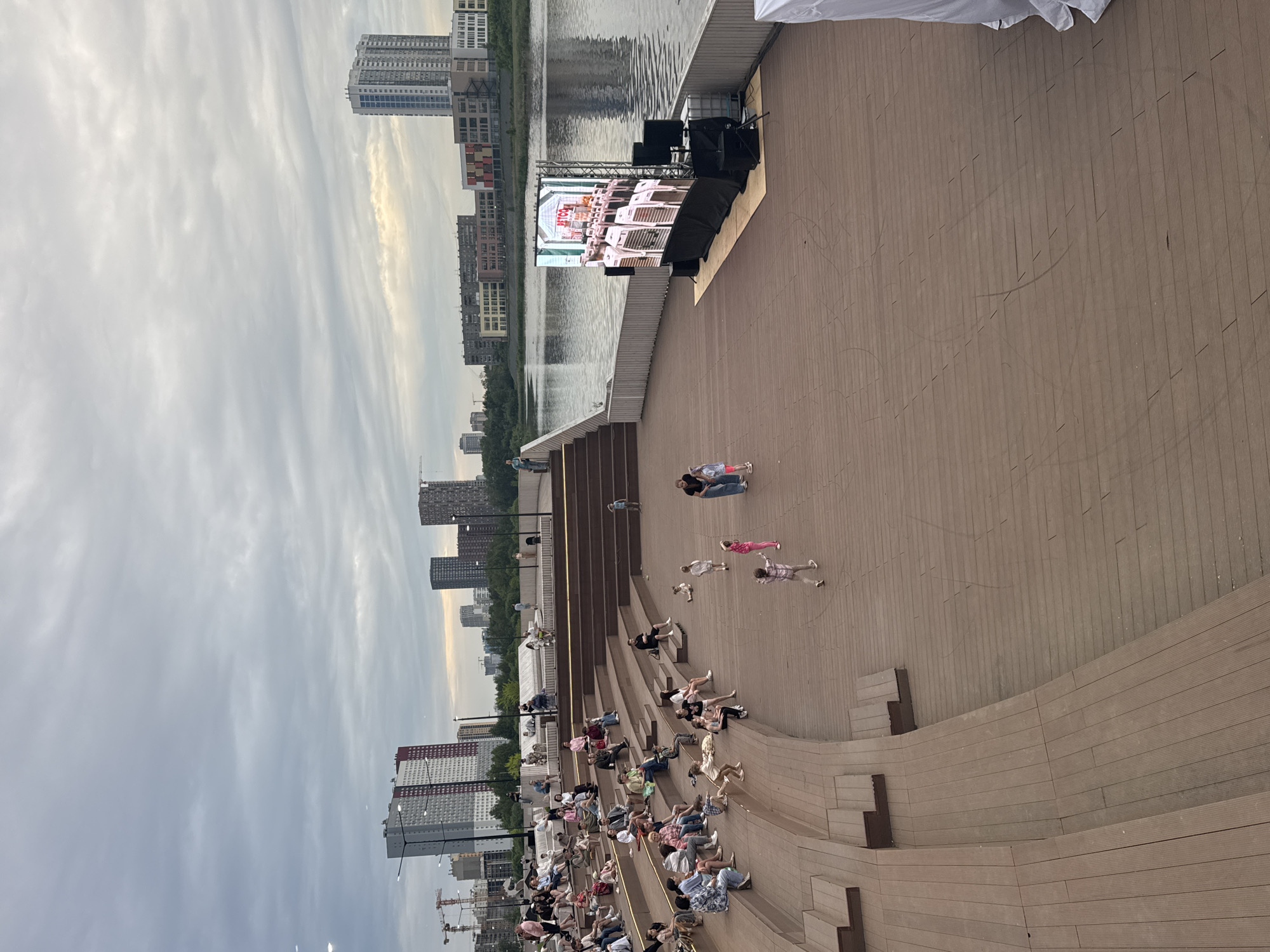 An outdoor amphitheater with tiered wooden seating overlooks a body of water. Many people are seated in the amphitheater, while others are standing on the wooden deck in front of it. A large LED screen displays an image of a Petco store interior. In the background, a cityscape with modern buildings is visible. The sky is overcast. The location is the "Ural-100" amphitheater in Yekaterinburg, Russia.
