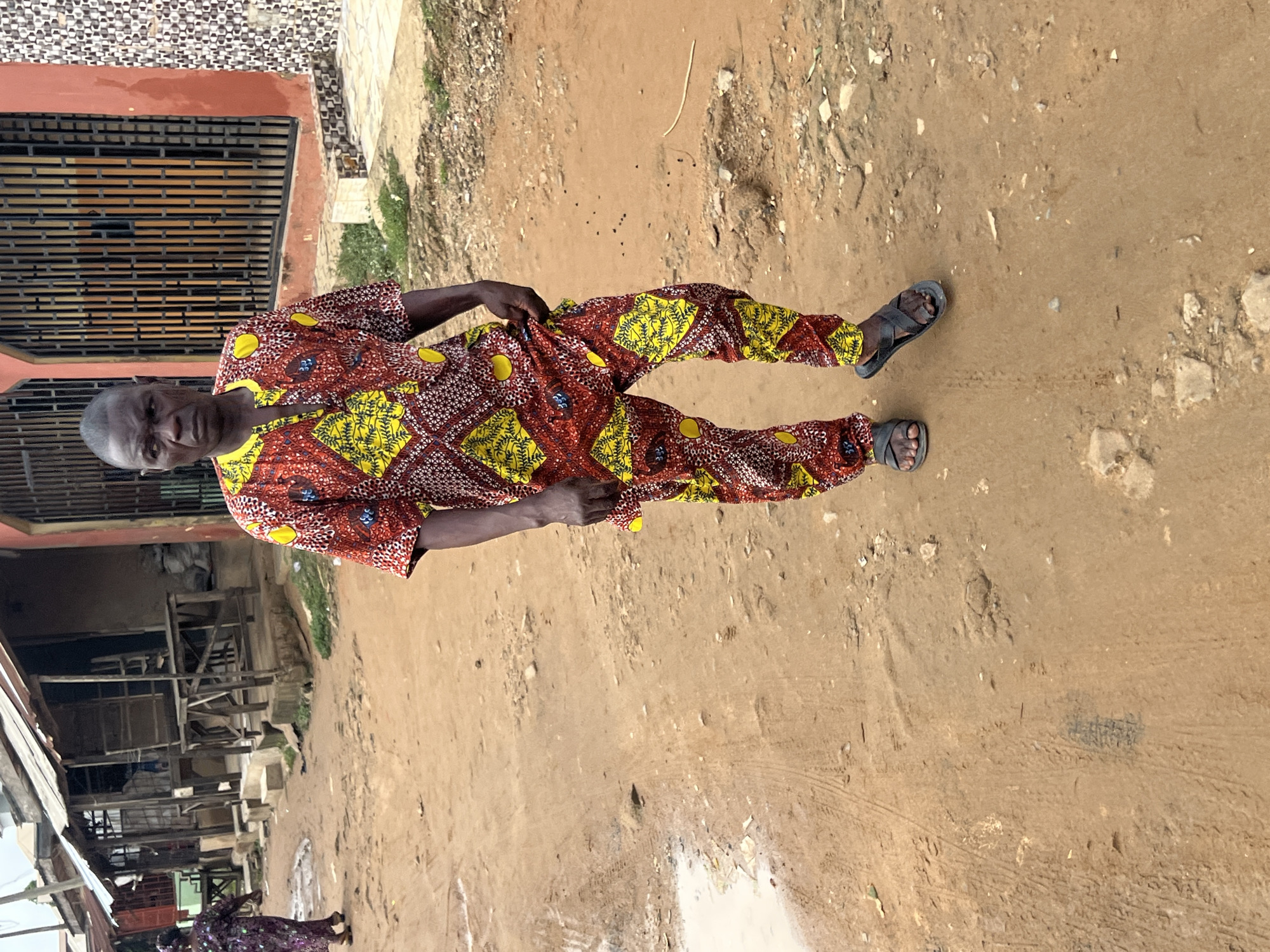 A man stands in a dirt street in Ijegun, Nigeria. He wears a patterned shirt and pants, and sandals. Behind him is a building with barred windows. To the left is a shop. The street is wet.