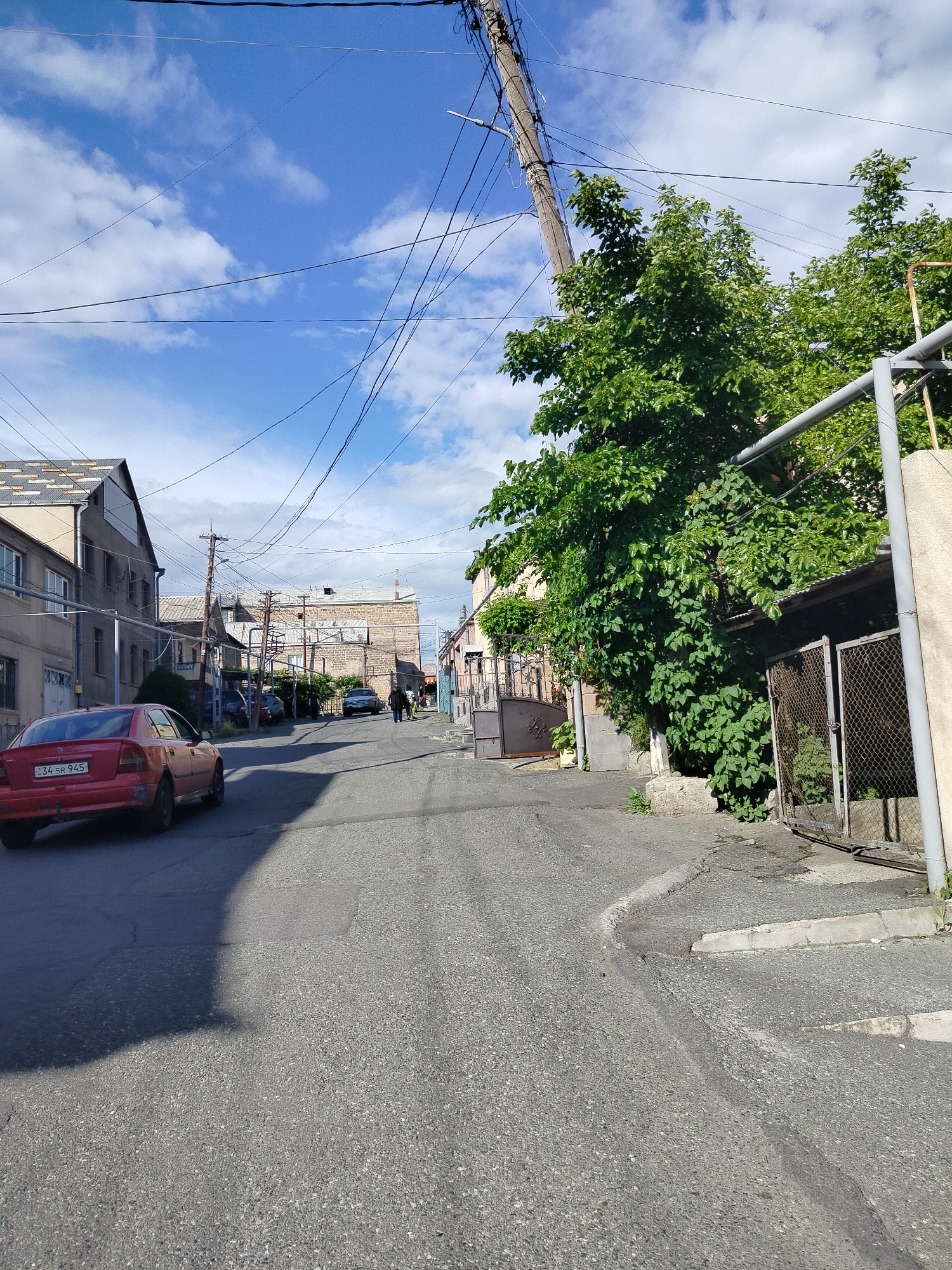 The image depicts a street scene in Yerevan, Armenia. The road slopes upward, with a red car parked on the left side, partially casting a shadow. Buildings line both sides of the street. Several utility poles and power lines crisscross overhead against a blue sky with scattered clouds. Two figures are walking uphill in the distance. Lush green foliage is visible on the right side of the street, obscuring some of the building facades. A chain-link fence structure stands to the right, adjacent to the sidewalk. The license plate on the red car is visible, reading "34 SR 945".
