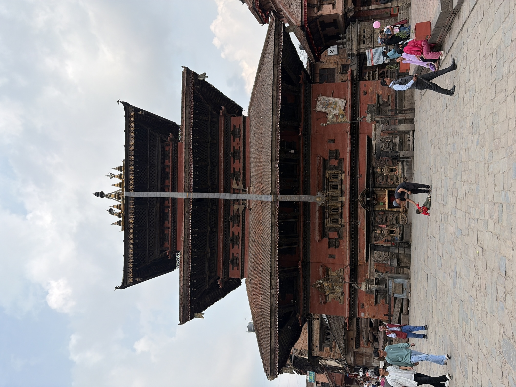 This vertical image depicts a bustling scene in what appears to be a historic town square, specifically a Durbar Square in Bhaktapur, Nepal. The dominant subject is a majestic, multi-tiered pagoda-style temple, characterized by its red brick base, intricately carved wooden elements, and stacked roofs culminating in golden finials (gajur) at the top. A tall, slender pillar stands prominently in front of the temple.
The paved stone square in the foreground and to the right is populated by several individuals. People are seen walking in various directions; for instance, a man in a light blue sweatshirt and a woman in a white top are walking towards the right side of the frame. Towards the far right, closer to the temple's base, a man in a waistcoat and white shirt stands, while nearby, a woman in pink holds a pink balloon. Further back, near the temple entrance, a person in a dark cap interacts with a child in a red jacket. The general activity suggests a regular day with people going about their daily routines or visiting the site.
The setting includes other traditional red-brick buildings with detailed wooden windows and balconies adjoining the main temple structure. The time of day is daytime, under a partly cloudy sky, indicating bright ambient light. There are no strong emotions evident among the people. Visible text on signs attached to the buildings is present, but too indistinct to be read clearly. The architecture is distinctive of the Kathmandu Valley, consistent with the location context of Bhaktapur.