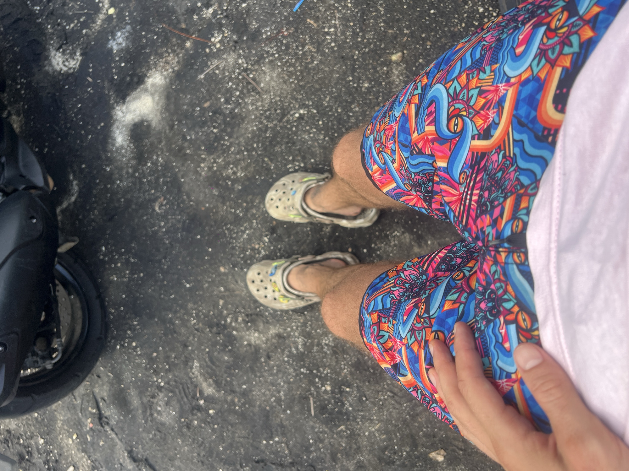 An overhead, first-person shot captures a person standing outdoors in Selemadeg, Indonesia. The subject's legs are visible, dressed in vibrant, multi-colored shorts featuring an intricate psychedelic pattern with elements resembling eyes, waves, and abstract floral or geometric shapes in various shades of blue, pink, orange, and red. They are wearing light grey Crocs on their feet. A portion of a light-colored (possibly white or light pink) t-shirt and the person's right hand resting on their thigh are visible on the right side of the frame.
The ground beneath is a dark, gritty surface, likely asphalt or a paved area, speckled with numerous small, light-colored particles, possibly sand or dust, and a few larger white patches. In the bottom-left corner, the front wheel and part of the body of a black motorcycle or scooter are visible, suggesting the person is either near or operating the vehicle. The scene is well-lit, indicating it is daytime. The casual attire and the presence of a scooter are common in the tropical climate of Indonesia. No specific activity or emotions are discernible, and no text is visible in the image.