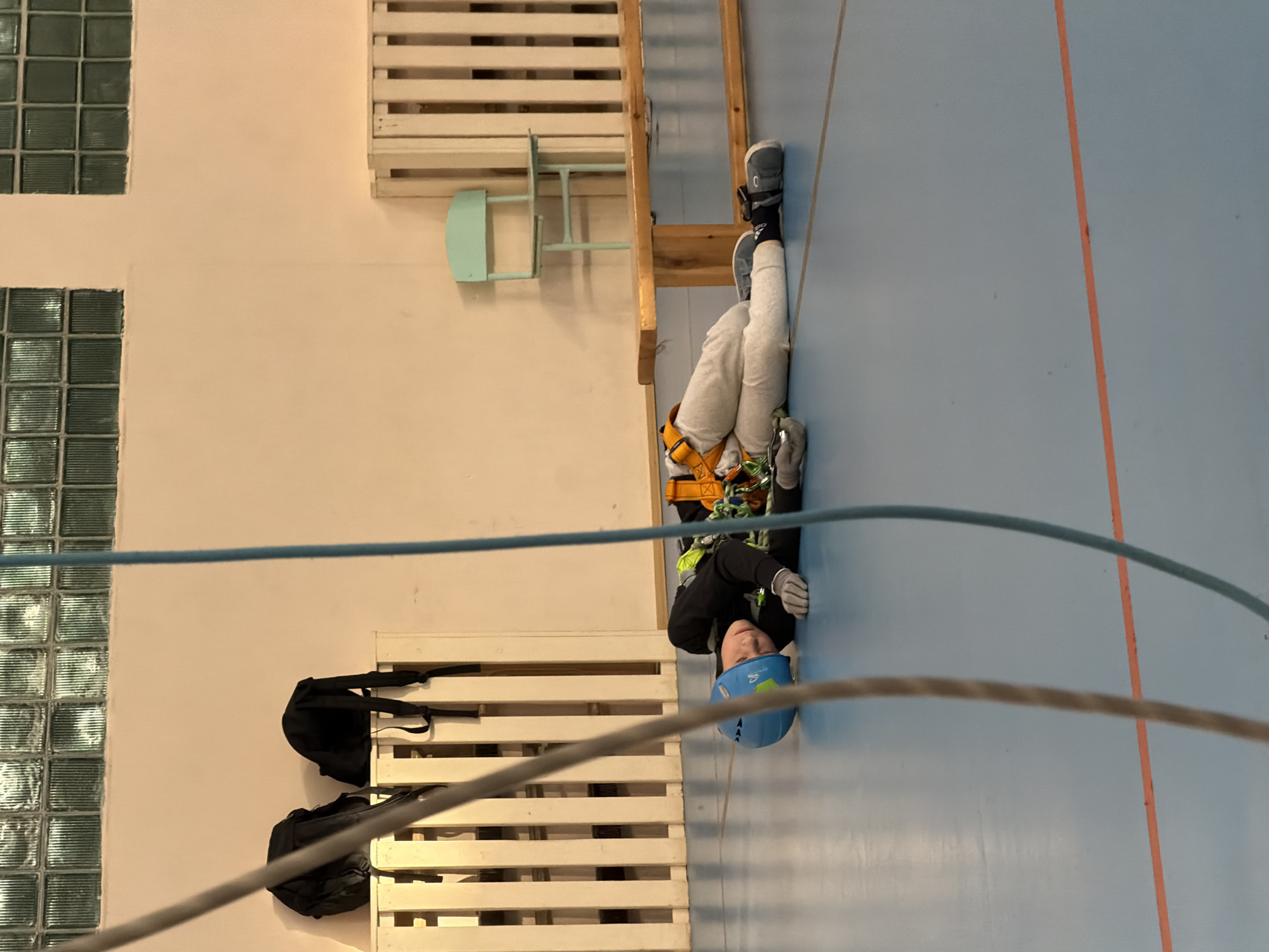 A person wearing a blue helmet, grey gloves, black long sleeve shirt, grey sweatpants, and grey shoes with a yellow harness is suspended from a wooden structure with a grey cable. The location appears to be indoors, possibly a gym or training facility, given the blue floor with a single orange line. Other objects visible are a pale green chair, a white wooden structure, two black bags on a white wooden structure and glass block windows. The city of Berezniki, Russia is not discernible.