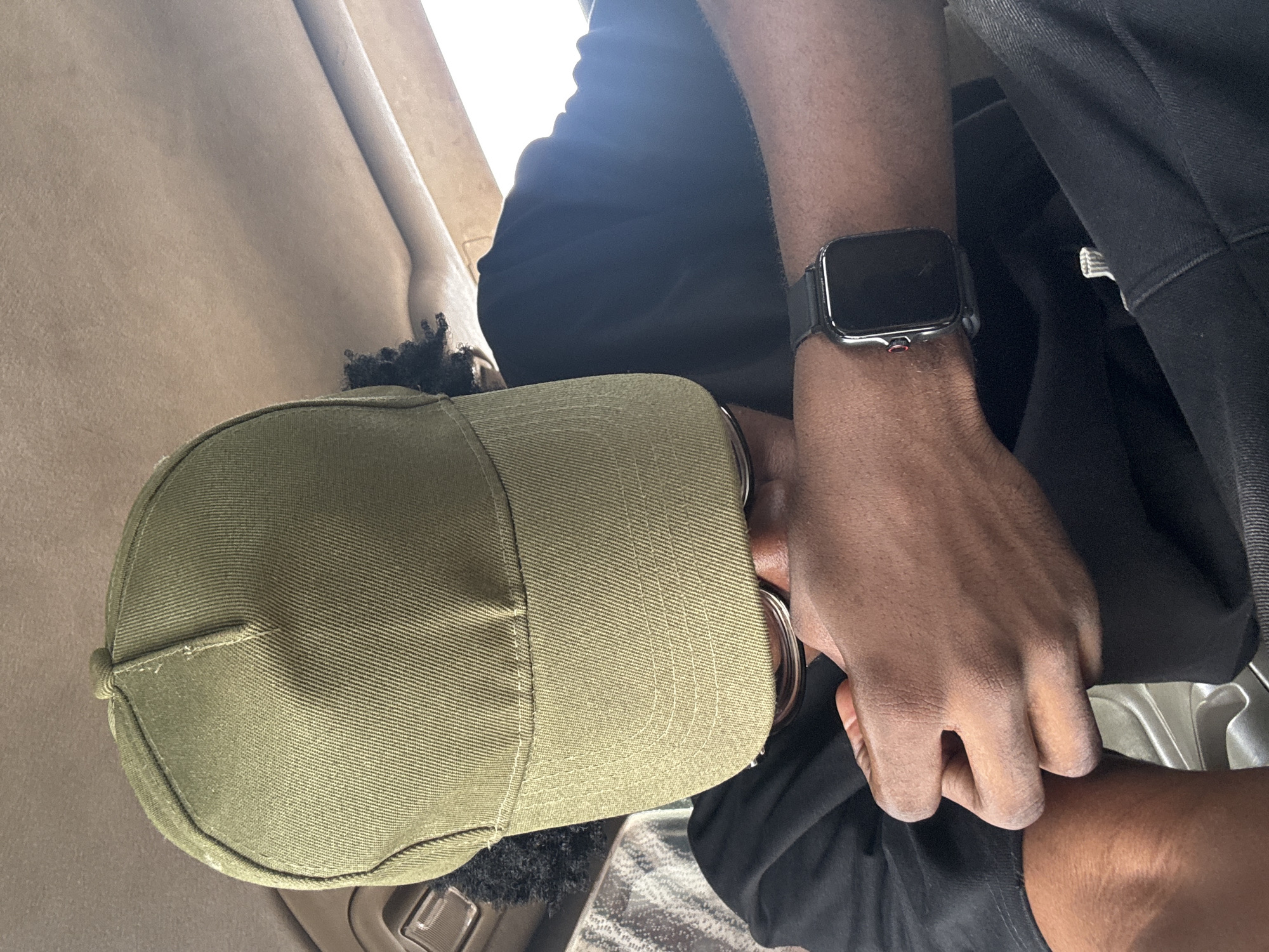 The image shows a person from the chest up, wearing a green baseball cap and a black t-shirt. They are also wearing a smartwatch on their left wrist. The person's hands are visible, and one hand appears to be resting on their lap. The background suggests an indoor setting, possibly a car or a room with light-colored upholstery. The lighting indicates it is daytime, with sunlight streaming in from the side. The scene is a close-up shot, focusing on the person's attire and accessories. There is no visible text in the image. The location context provided is Orji, Nigeria.