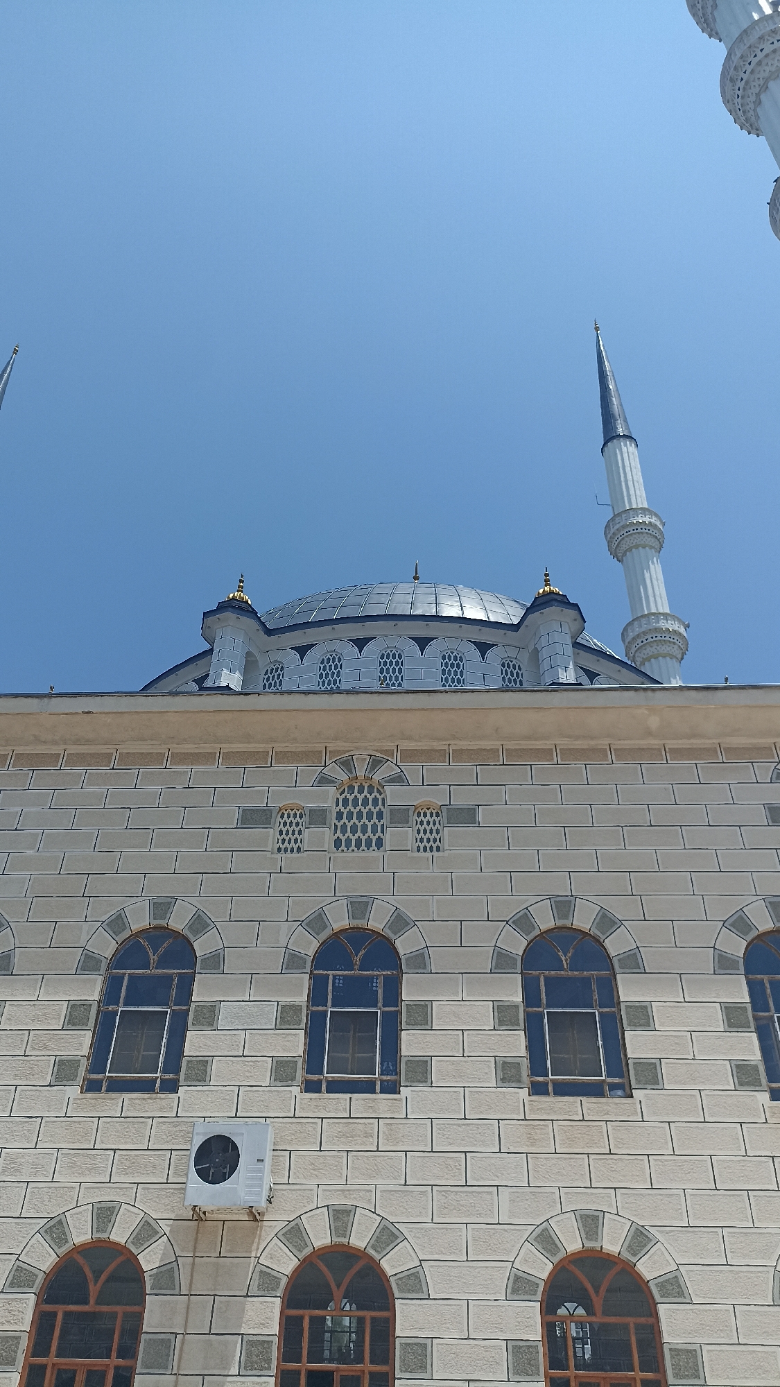 A low-angle, close-up shot shows the exterior of a mosque, with a clear blue sky in the background. The mosque features a large, domed roof with a decorative finial, and a tall, slender minaret with a pointed top. The building's facade is constructed of light-colored stone blocks arranged in a brick pattern, with arched windows featuring intricate latticework. Some of the windows are large and rectangular, while others are smaller and set within arched recesses. An outdoor air conditioning unit is mounted on the wall below a row of windows. The overall impression is of a modern, well-maintained religious structure.