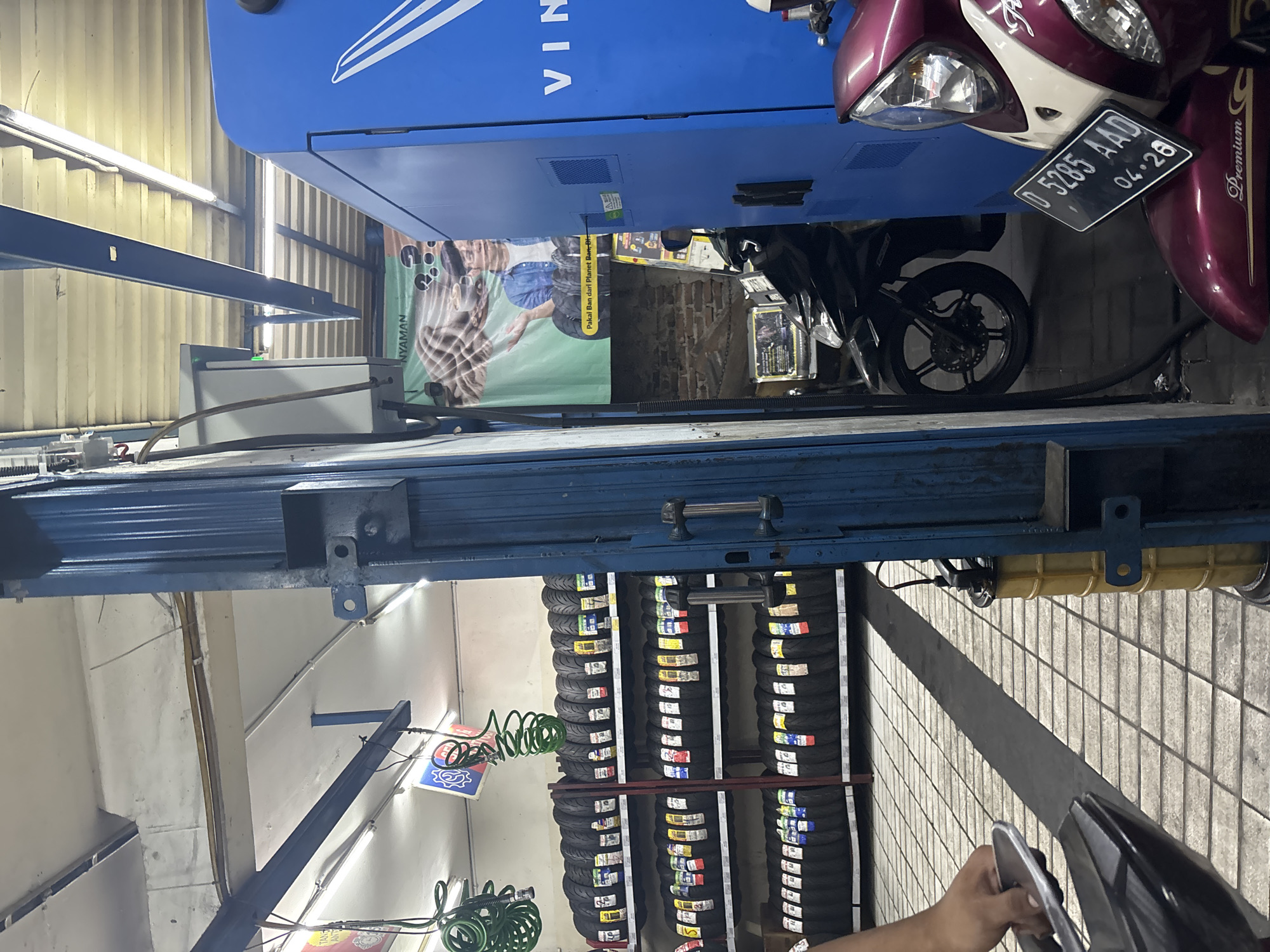 The image depicts the interior of a tire shop in North Jakarta, Indonesia. Multiple shelves are stacked with tires, ranging in size and brand, suggesting a commercial setting for vehicle maintenance. A blue dispensing machine, possibly for fuel or air, is situated to the right of the frame, branded with "VIN". In the foreground, the front section of a burgundy scooter is visible, displaying a license plate with the number "D 5285 AAD" and the date "04.28". Another black motorcycle is parked behind the scooter. A person's hand is visible on the left, holding what appears to be a tire-related tool or accessory. A banner with an image of a man and text in Indonesian hangs on the wall behind the dispensing machine. The floor is tiled in a grid pattern, and fluorescent lighting illuminates the space.