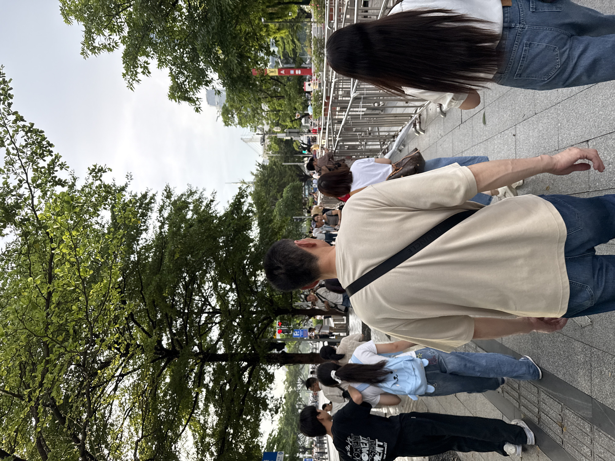 In Haizhu District, China, a group of people walks along a paved sidewalk lined with trees and metal barriers. The sky is overcast. In the foreground, a man wearing a beige t-shirt and jeans walks away from the camera. To his left, a person in a black t-shirt and dark pants walks alongside another person in a white t-shirt and jeans, who carries a light blue backpack with a cartoon face. Further ahead, other individuals are seen walking in the same direction, some obscured by the crowd and the metallic barriers. Traffic lights are visible in the background, indicating a street intersection. The environment appears to be a public urban area, possibly near a park or a busy street.