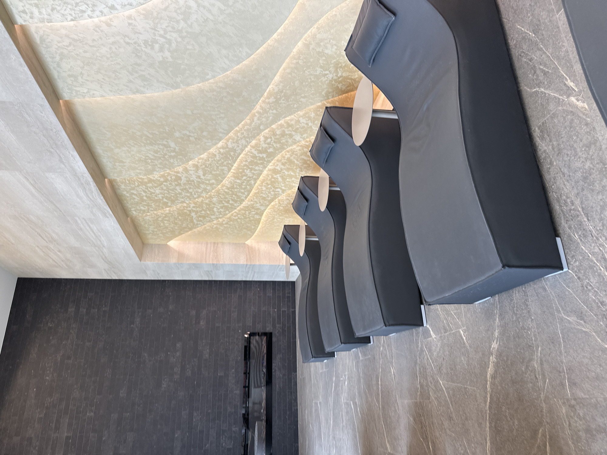 A low-angle interior shot depicts modern lounge chairs set against a textured wall. The room features unique ceiling designs with wavy, illuminated patterns. The chairs are upholstered in a dark gray or black material, arranged in a line, giving the impression of multiple seating options in a waiting area. A rectangular mirror or display is visible on the adjacent wall. The specific location within Kazanlak, Bulgaria, cannot be determined from the image.