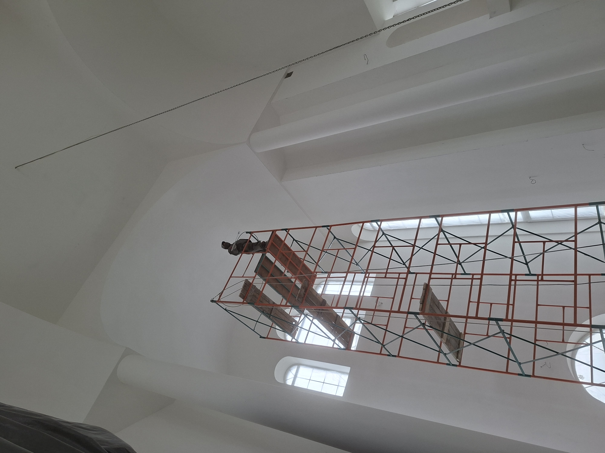 The image shows the interior of a building, possibly a church or similar structure, in an unknown location. The dominant color is white, used extensively on the walls and ceiling. The ceiling has a multi-tiered design with sections protruding downward. A series of windows are visible. There is a large orange scaffolding-like structure with green supports hanging from the ceiling, on which rests what seems to be a wooden effigy of a religious figure or a simple cross. The scaffolding is located between the camera and the windows and casts a shadow on the ceiling. A thin chain-like cable stretches across the top part of the ceiling.