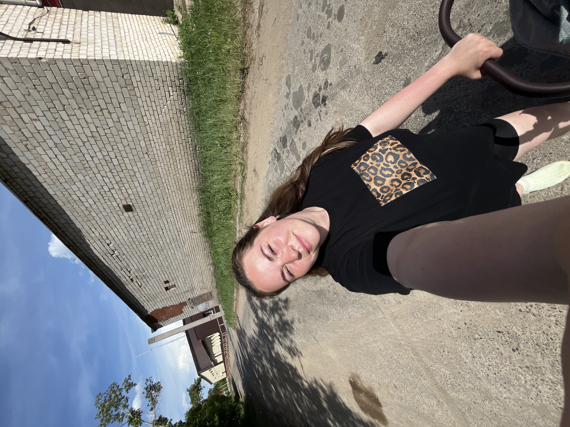 A woman is seen from a low angle, possibly a selfie, standing on a cracked asphalt surface next to a building. The building has a brick facade and is located near a grassy area. In the distance, there are other buildings and trees. The sky is visible above. The woman has long brown hair and wears a black shirt with an animal print detail. She appears to be holding onto a circular object, possibly a handle. The photo might be taken in or near the city of Chaikovsky, Russia, if specified as such by external information.