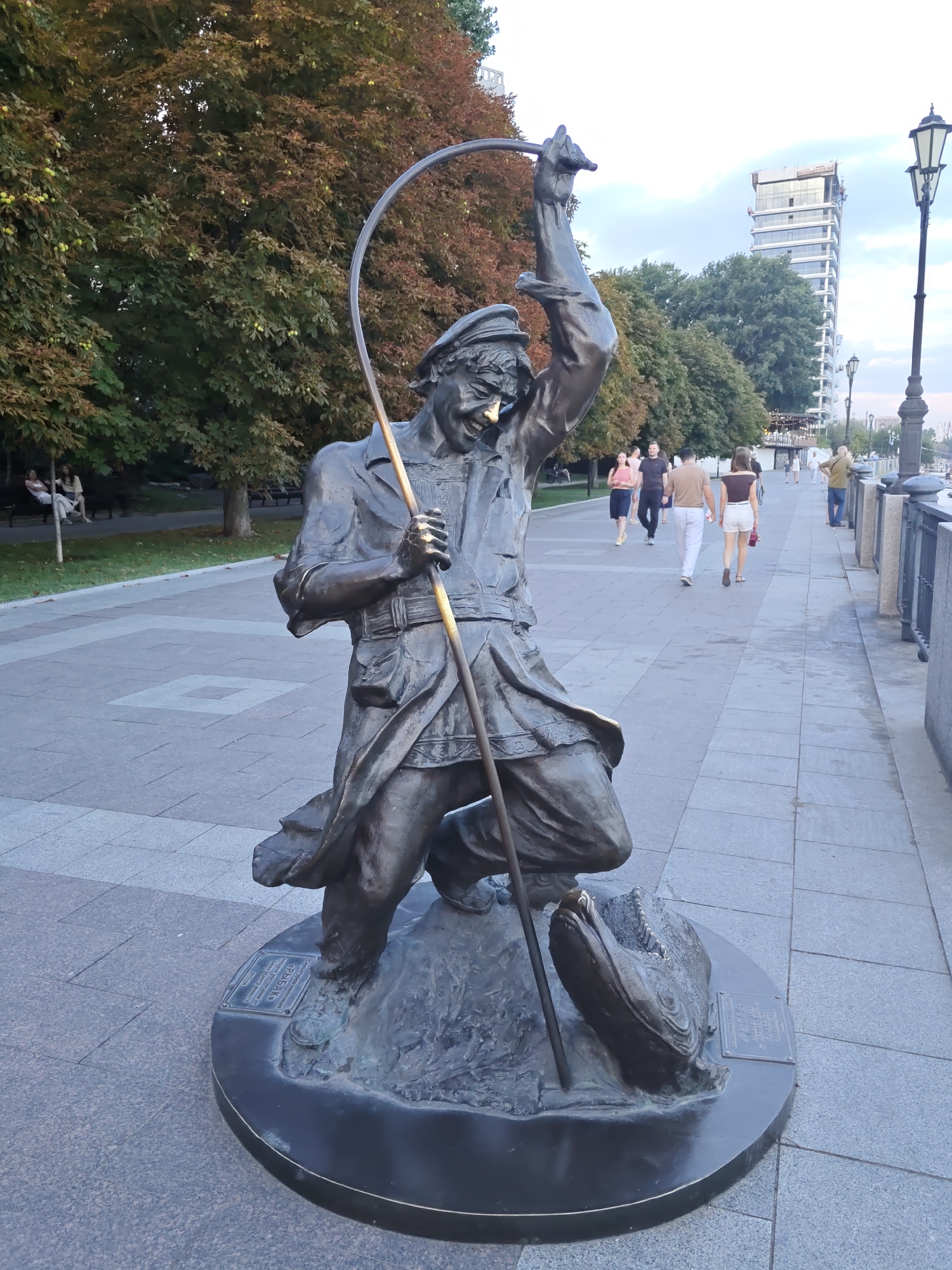 A bronze statue of a fisherman stands on a circular bronze base on a paved walkway. The fisherman, depicted with a large smile and a triumphant pose, holds a fishing rod curved over his head. He is dressed in a cap, jacket, and trousers. At the base of the statue, a large fish is partially visible. The walkway is lined with trees on the left and a river embankment with a railing on the right. In the background, there are several people walking along the path and a tall, modern building under construction. The sky is overcast. This statue is located on the Don Embankment in Rostov-on-Don, Russia.