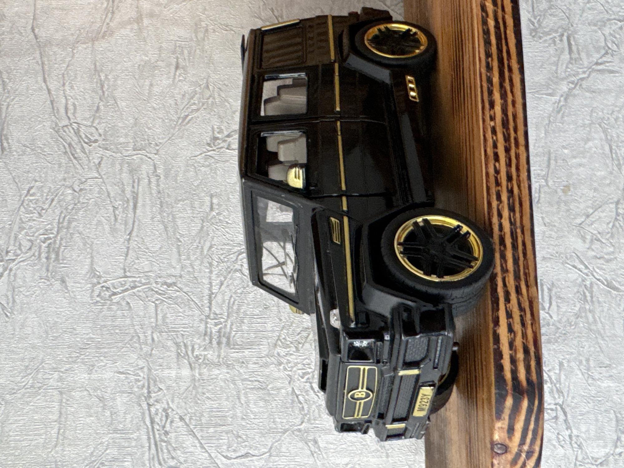 A black die-cast Mercedes-Benz G-Wagon toy car with gold accents sits on a wooden shelf against a textured white wall. The car has a license plate that reads "M923Y". The interior is visible and appears to be tan. The car is detailed with gold trim on the grille, side mirrors, door handles, and wheels. The wall behind the car has a crinkled, almost crumpled texture. The wooden shelf has a visible wood grain pattern and appears to be part of a larger piece of furniture. There are no people or animals present in the image. The scene is indoors, possibly in a home or display setting. The location of Яромінскі сельскі Савет, Belarus is not visually represented in this image.