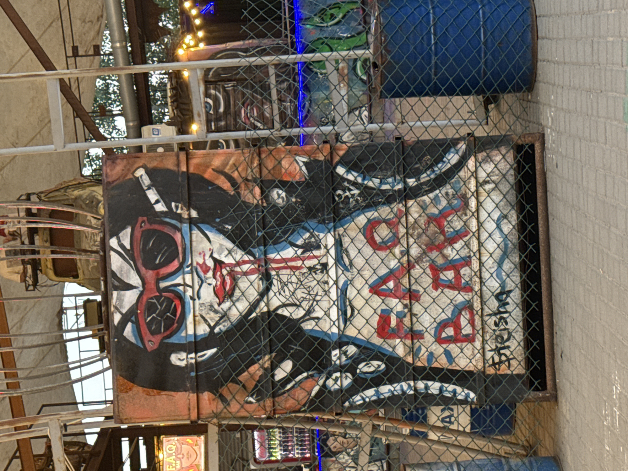The image captures a vibrant street art mural painted on a corrugated metal surface, possibly a repurposed oil drum or panel, located in the city of Yarovoye, Russia. The artwork features a stylized depiction of a person with striking red sunglasses, dark hair, and red lips with red streaks resembling blood. Below this portrait, the words "FAQ BAR" are spray-painted in red and blue letters, with smaller, handwritten text that appears to read "steisha" underneath. The mural is partially obscured by a chain-link fence. In the background, an assortment of objects hints at a lively outdoor setting: a ladder leans against a structure, decorative lights twinkle, and several blue oil drums are visible. The overall impression is one of an eclectic and artistic urban space.