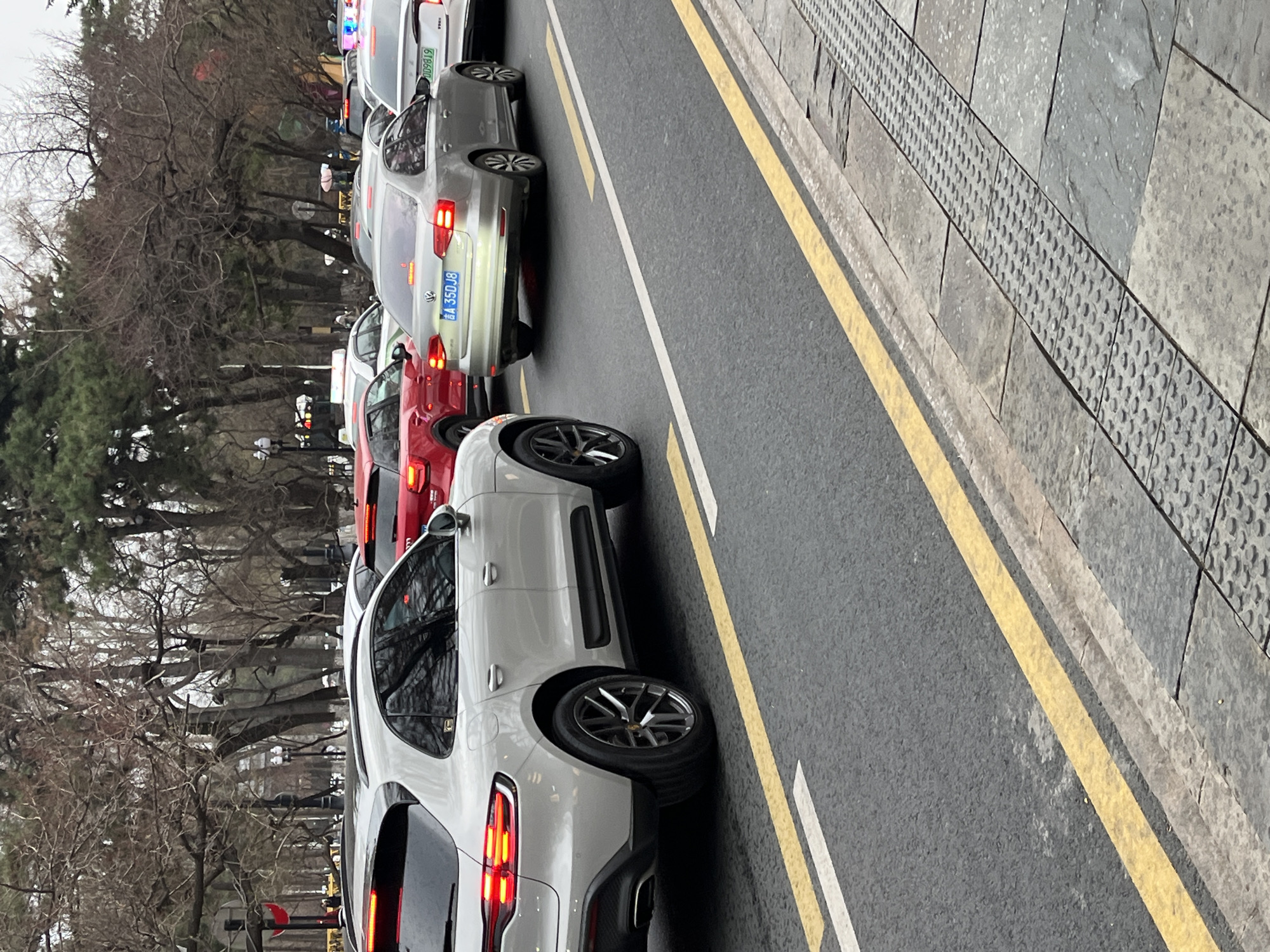The image depicts a congested street in Changchun City, China. Multiple cars, including a white SUV, a red sedan, and a silver sedan are lined up bumper-to-bumper in traffic. The vehicles are positioned on an asphalt road marked with yellow and white lane dividers. Alongside the road is a grey sidewalk with a textured, non-slip surface. Bare trees without leaves are visible on the left side of the frame, suggesting a winter or early spring season. The presence of license plates indicates the cars are operational vehicles. The overall scene reflects urban traffic conditions.