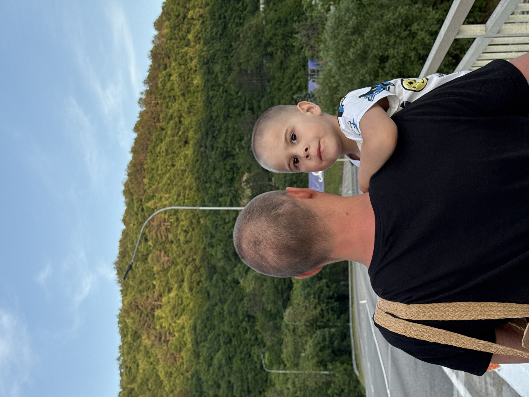 The image depicts a man carrying a young boy on his right shoulder, standing on what appears to be a bridge or overpass, in Nebug, Russia. The man is seen from the back, with the top of his head and shoulders visible. He has short, light brown hair and is wearing a black t-shirt. A light brown, woven strap bag is visible on his left shoulder. The young boy, facing towards the viewer, looks directly at the camera with a neutral expression. He has short, light brown hair and is wearing a white t-shirt with blue and yellow patterns, including a yellow smiley face. His left arm is resting on the man's shoulder.
Behind the figures, a paved road with white lane markings is visible to the left. A metal guardrail extends along the right side of the bridge. In the background, a dense, forested hillside rises, covered predominantly in green deciduous trees, with some interspersed lighter brown foliage suggesting evergreens or a different species. The sunlight illuminates the trees from the right, creating a distinct play of light and shadow. Two streetlights with curved arms are visible, one prominently arching from the left and another further back on the right. The sky above is clear and blue with sparse, wispy white clouds, indicating late afternoon or early evening.