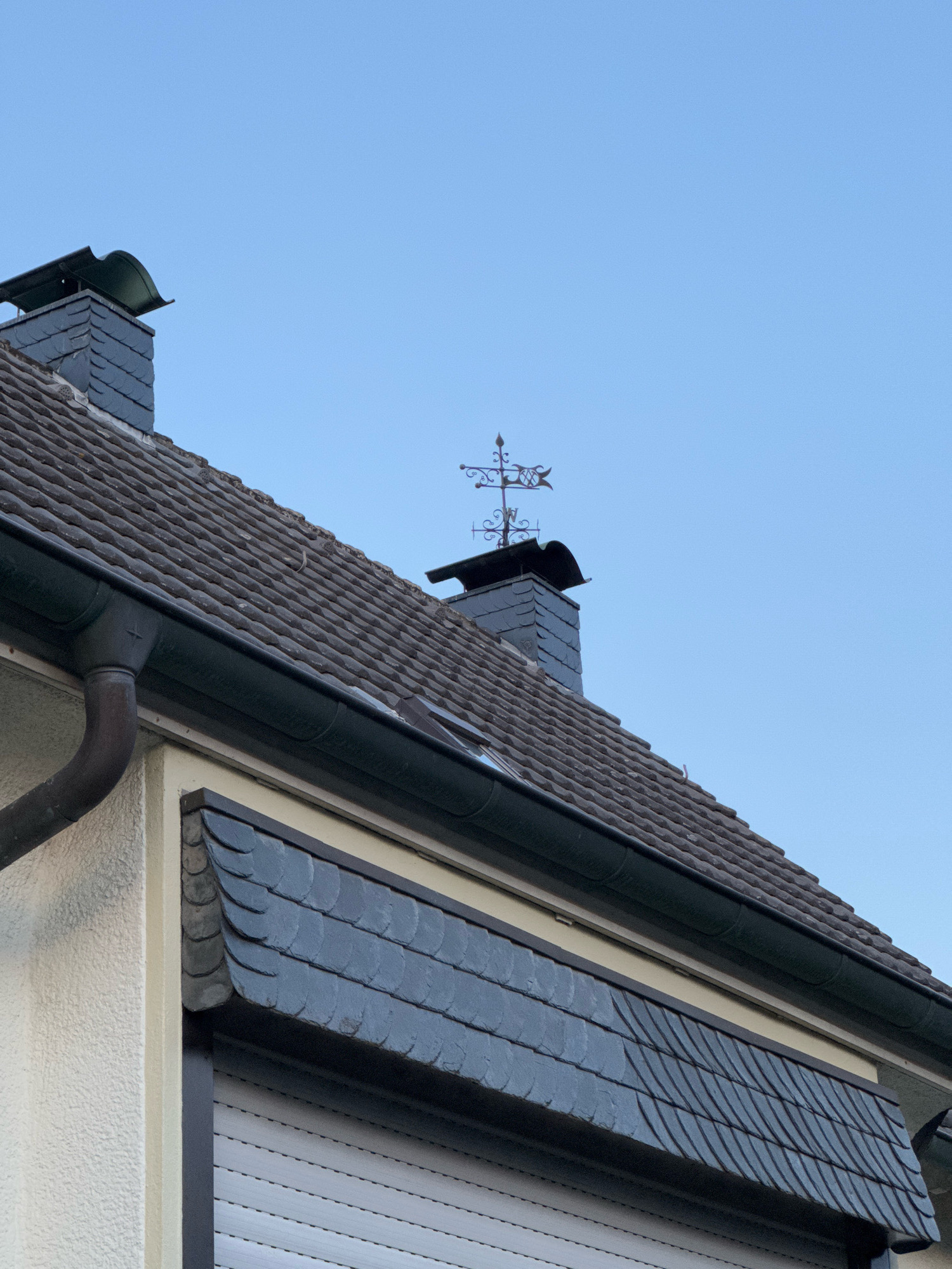 This is a low-angle, eye-level exterior shot of a house in Cleves, Germany, on a clear day with a light blue sky. The roof is covered in dark gray, textured tiles. To the left, a brown downspout is attached to a dark gray gutter. A chimney, also clad in dark gray tiles, rises from the roof, topped with a decorative weather vane. Below the roofline, a textured, light-colored wall is visible, with a dark gray awning composed of overlapping slate tiles extending over a grey rolling shutter. The image does not contain any people or indicate any specific actions or interactions. The scene depicts a typical residential architectural element in a German town.