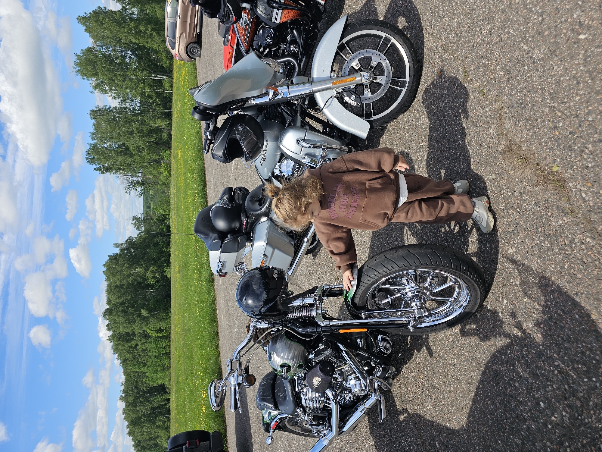 The image shows a child in a brown tracksuit standing next to a black and chrome motorcycle on an asphalt surface. The motorcycle is equipped with a skull-themed fuel tank design and a black helmet rests on the seat. Two other motorcycles are parked nearby, including a silver one. In the background, there's a green field bordering a tree line beneath a blue sky with scattered clouds. The location is presumably near Borodinskogo Muzeya, Russia.