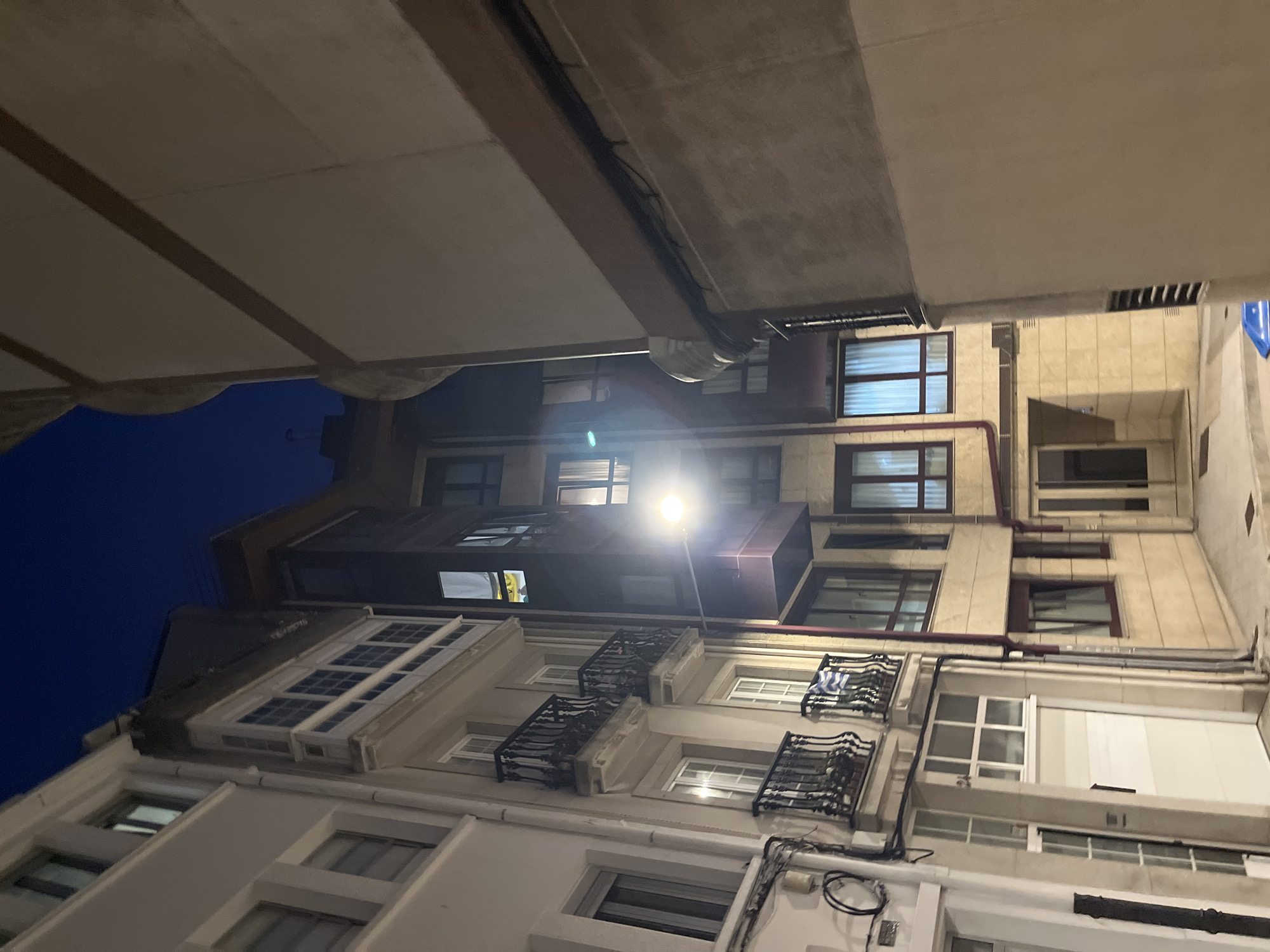 The image shows a narrow urban street in A Coruña, Spain, taken from a low angle looking up at the buildings. The buildings are closely packed together and appear to be multi-storied, with a variety of window styles and sizes. Some windows have wrought-iron balconies. There are drainpipes running down the sides of some buildings. Overhead, there is a concrete or plaster structure, partially obscuring the sky. The sky is a dark blue, suggesting it is nighttime or early morning. A streetlight is visible, providing illumination and casting light onto the building facades. Numerous wires are strung across the front of the buildings. The buildings are primarily white and tan.