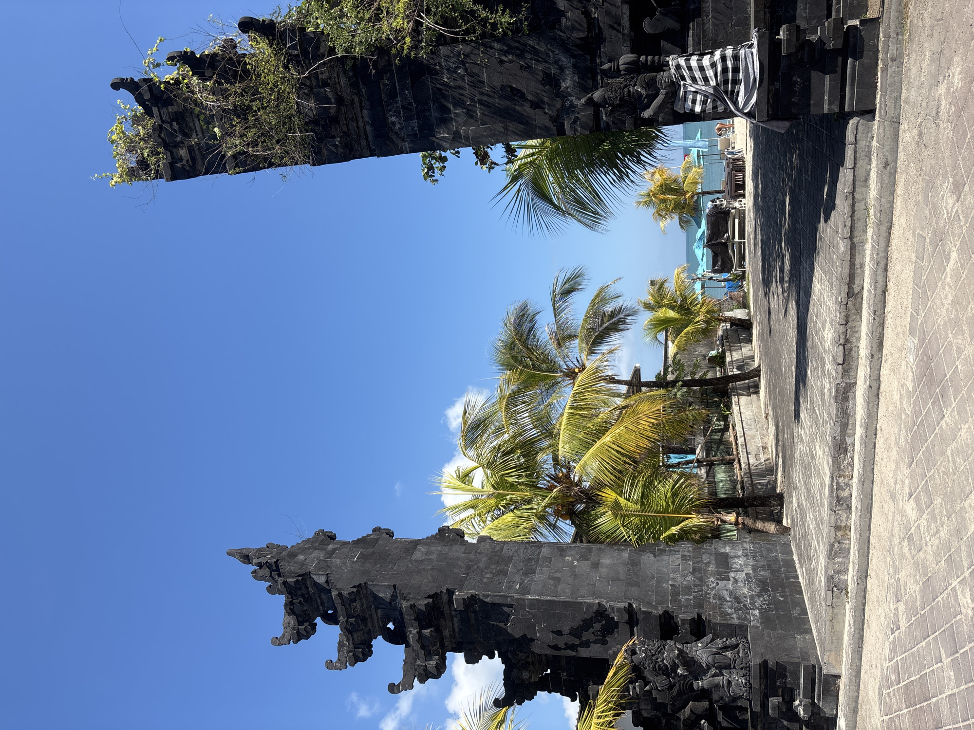 The image depicts a traditional Balinese stone gate in Kuta, Indonesia, framing a view of a tropical beach. The gate, made of dark volcanic stone, features intricate carvings and is adorned with overgrown vegetation on the right pillar. Through the archway, palm trees with bright green fronds line a paved walkway leading to a beach. Beach chairs and umbrellas, in shades of blue and white, are set up on the sandy shore, and the ocean stretches out to the horizon under a clear blue sky with scattered white clouds. A person is visible near the water, and a patterned textile is draped over a statue on the right side of the gate. The foreground is paved with stone tiles.