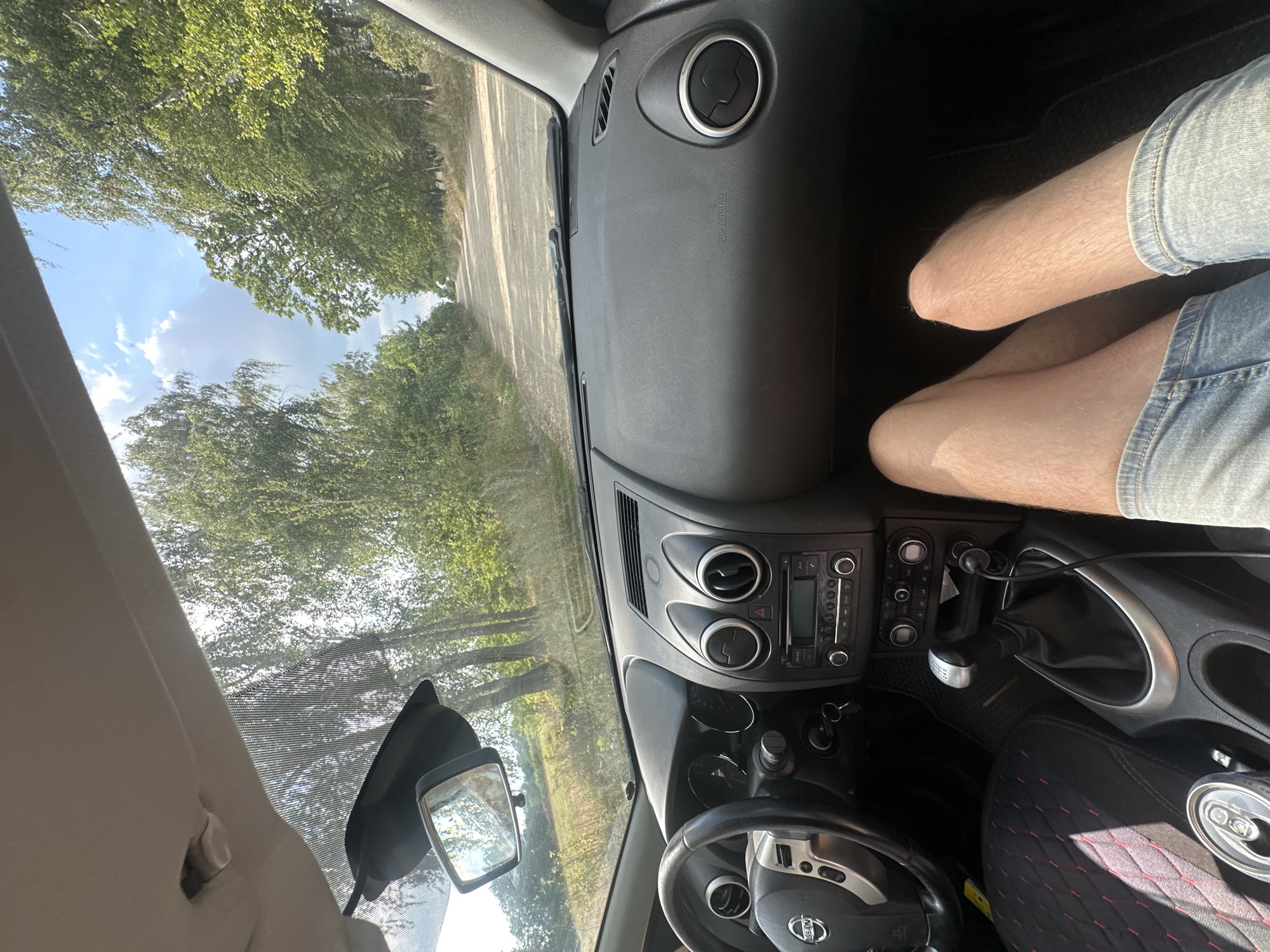 The image shows the interior of a car, looking forward through the windshield. The dashboard and steering wheel are visible on the left. A person's legs, wearing denim shorts, are in the foreground. Outside the car, a dirt road is visible, flanked by trees and greenery, under a partly cloudy sky. The scene is set on a sunny day. The location is Tynna, Ukraine.