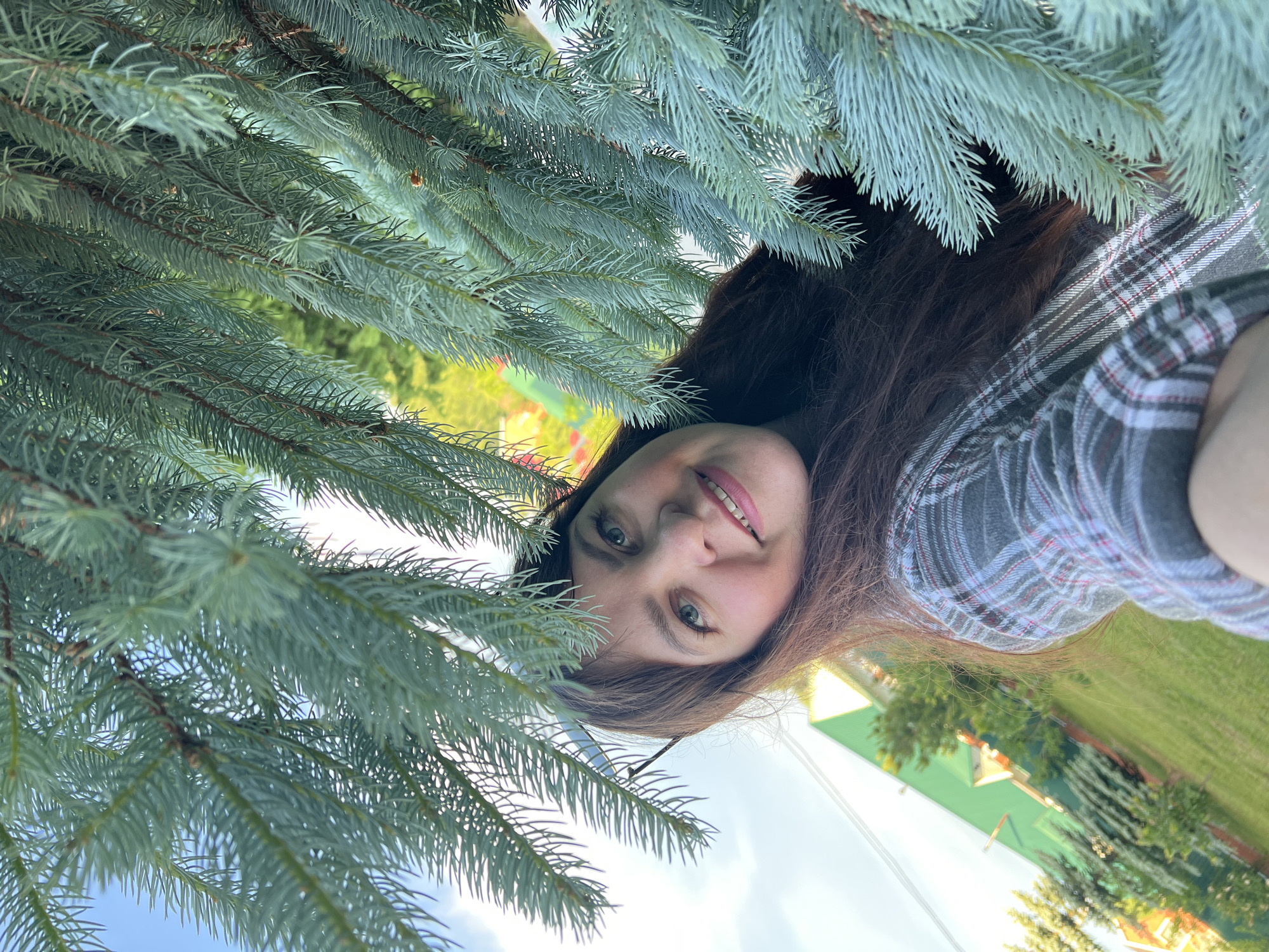 An outdoor portrait shot in Noviy, Russia, shows a woman's face peeking through the dense branches of an evergreen tree, likely a blue spruce, which fills the foreground. The image is rotated ninety degrees, making the woman appear to be looking upwards from beneath the branches.
The subject is a young to middle-aged woman with long, dark brown hair and clear blue-green eyes. She has a gentle, inviting smile, revealing her teeth, and her expression is friendly and engaging. She appears to be wearing a plaid shirt with shades of grey, white, and red. Her face is in focus, while the background and some of the immediate foreground branches are slightly blurred, creating depth.
Behind the woman and the tree branches, the background reveals a bright, sunny day with patches of clear blue sky. A vibrant green lawn or field is visible, along with distant trees and what appears to be a building with a distinctive bright green roof, possibly a house or outbuilding. The natural lighting suggests it is daytime, possibly morning or late afternoon, with soft, even illumination. The overall scene conveys a serene and pleasant outdoor environment. No visible text is present in the image.