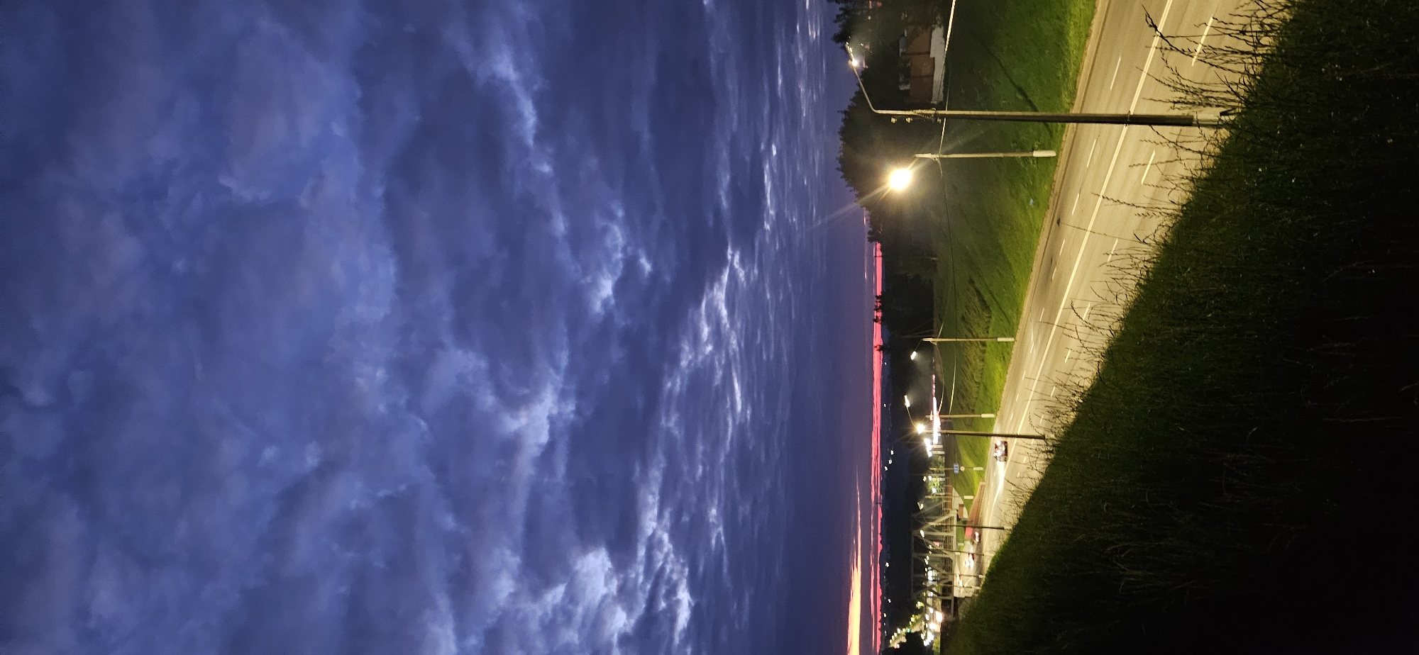 The image depicts a twilight scene on a road in Vitsebsk, Belarus. The sky is filled with dark, textured clouds tinged with purple and hints of pink near the horizon. A highway, illuminated by streetlights, curves through a grassy embankment on the right. A few cars are visible on the road, their headlights on, indicating low visibility. In the distance, a bridge and buildings are faintly discernible, along with the silhouettes of trees. The overall impression is of a quiet, perhaps slightly melancholic, evening commute.
