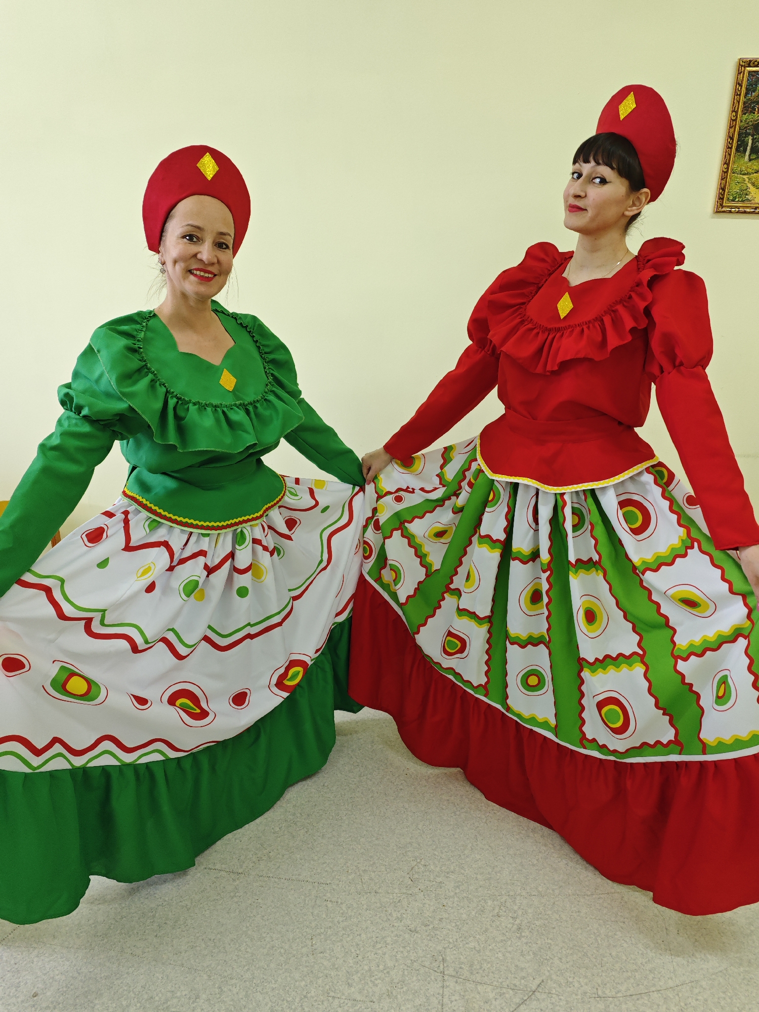 Two women are standing side-by-side indoors, posing in colorful folk-style costumes. The setting appears to be a brightly lit room with plain, light-colored walls and a light-colored, possibly tiled or linoleum, floor. A framed landscape painting is partially visible on the wall behind the woman on the right.
Both women are dressed in vibrant costumes featuring red and green. The woman on the left wears a bright green blouse with ruffled details around the neckline and long sleeves. Her outfit is completed with a skirt that has a green base layer, topped with a white overlay patterned with red, green, and yellow wavy lines and abstract circular and oval shapes. She wears a red headpiece adorned with a yellow diamond accent, and a matching yellow diamond is on her blouse. She smiles directly at the camera.
The woman on the right wears a bright red blouse, also with ruffled details on the neckline and long sleeves. Her skirt similarly features a red base layer with a white patterned overlay that echoes the design of the other costume, showcasing red, green, and yellow shapes. She also wears a red headpiece with a yellow diamond accent, and a matching diamond is on her blouse. She looks slightly away from the camera with a subtle smirk.
Both women are holding out their skirts, possibly to showcase the patterns or as part of a dance pose. Their attire suggests they are involved in a cultural performance or event, possibly in Марковский, Russia, given the location context. There is no visible text in the image.