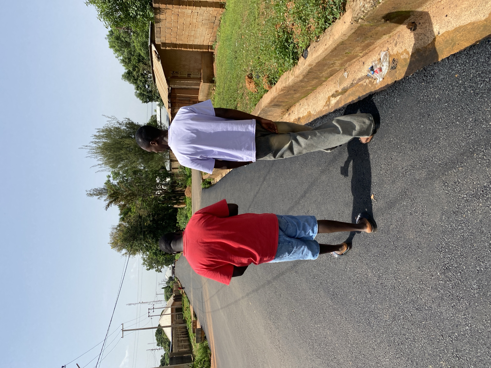 Two men are walking on a paved road in Bukuru, Nigeria. The man in front is wearing a red t-shirt and blue denim shorts. The man behind him is wearing a white t-shirt and light-colored pants. Both men are facing away from the camera. The setting appears to be a rural or semi-urban area, with buildings and vegetation visible in the background. The sky is clear and blue, suggesting it is daytime. There is a concrete gutter along the side of the road, with some trash in it. The road appears to be recently paved.