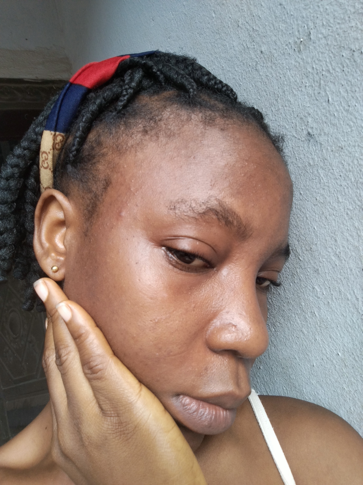 A close-up, right-facing profile shot shows a Black person with braided hair and a patterned headband that includes red, blue, and tan sections. A small gold stud earring is visible in the person's left ear. The person's right hand is resting on their face, with fingers near the cheekbone and chin. The background is a textured gray wall. The person is wearing a white strap, likely from a tank top or dress. The setting appears to be outdoors, against a wall, in Isiehor, Nigeria.