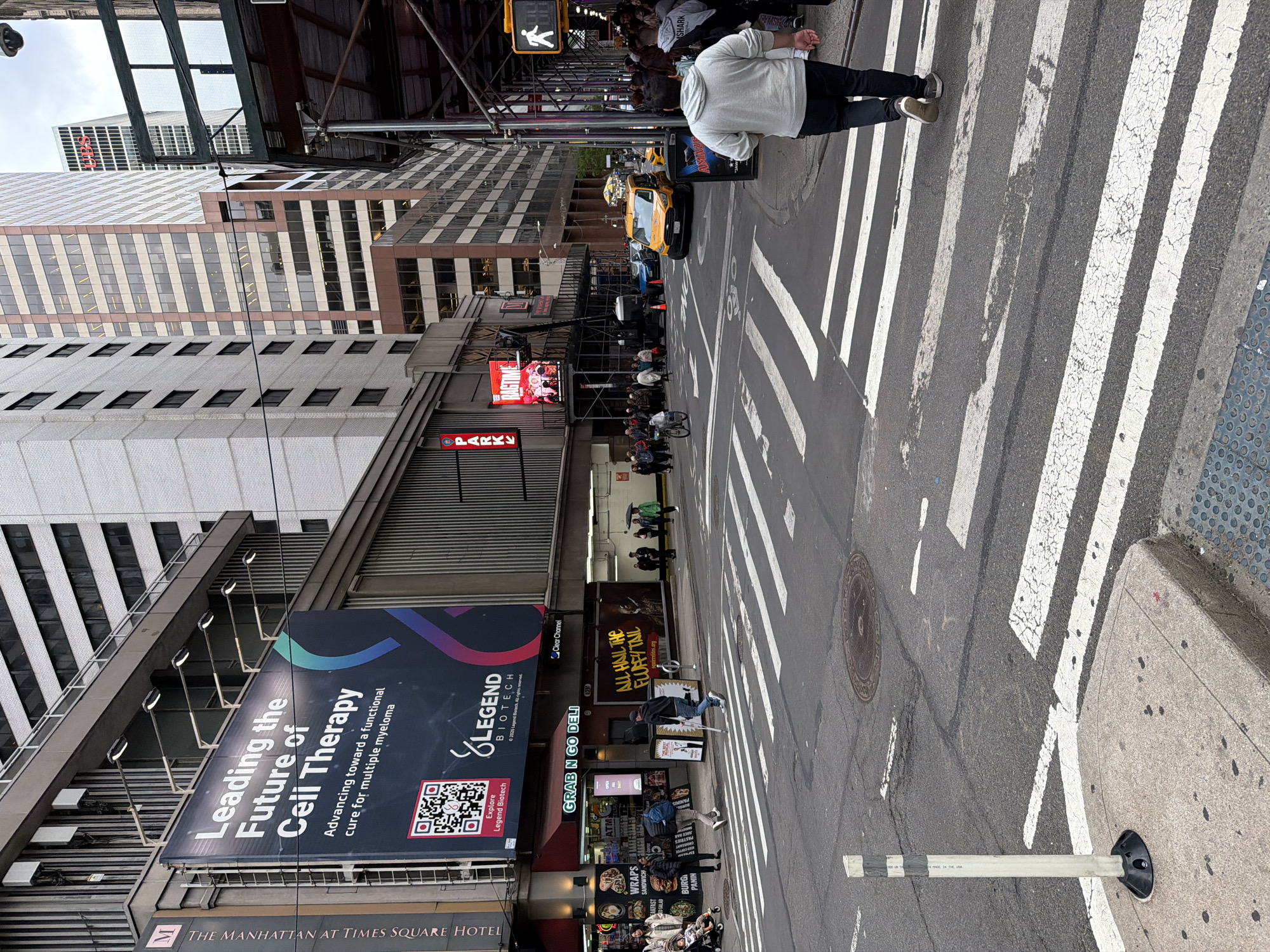 A daytime street scene in a bustling urban environment, presumably New York City, captures pedestrians, traffic, and towering buildings under an overcast sky. The image is taken from a low angle on a sidewalk, looking across a wide pedestrian crosswalk.
In the foreground, a person in a light grey hoodie and dark pants stands on the edge of a crosswalk, facing away from the viewer. The crosswalk's cracked white stripes extend across the asphalt street. A flexible white street bollard, marked "MADE IN THE USA," is visible near the curb.
Across the street, a yellow taxi cab is seen moving or parked amidst other vehicles and a flow of pedestrians. Many people are walking or gathered on the sidewalks. On the right, a "GRAB N GO DELI" entrance is visible, above which a digital sign displays "ALL HAIL THE FLUFFY TAIL" and "squirrelites.org." A red illuminated sign below reads "PARK." Overhead, dark metal structures suggest an elevated train line or building construction.
The left side of the frame is dominated by multiple high-rise buildings. A large vertical billboard is mounted on one, proclaiming "Leading the Future of Cell Therapy" and "Advancing toward a functional cure for multiple myeloma" for "LEGEND BIOTECH," complete with a QR code and copyright "©2025 Legend Biotech. All rights reserved." Part of the building's signage below reads "THE MANHATTAN AT TIMES SQUARE HOTEL," confirming the location context of New York City. In the distance, another skyscraper features a prominent red "SHARP" logo.
Visible text includes: "Leading the Future of Cell Therapy," "Advancing toward a functional cure for multiple myeloma," "Explore Legend Biotech," "LEGEND BIOTECH," "©2025 Legend Biotech. All rights reserved.," "THE MANHATTAN AT TIMES SQUARE HOTEL," "GRAB N GO DELI," "Clear Channel," "ALL HAIL THE FLUFFY TAIL," "squirrelites.org," "PARK" (multiple instances), "ATM," "WRAPS," "SANDWICH," "BREAKFAST," "HOT & COLD SALAD," "BURGER," "PANINI," "SHARP" (on a building), "GYMSHARK" (on a person's shirt), and "MADE IN THE USA" (on a bollard).