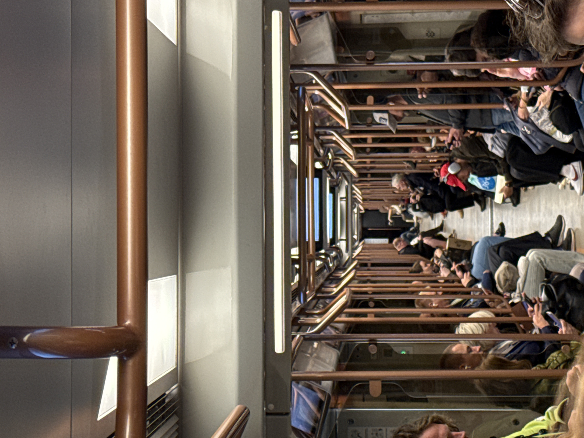 The image shows the interior of a Moscow Metro train car. Passengers are seated on both sides of the aisle, facing each other. Many are looking at their phones, and some are wearing headphones. The train car has a modern design with copper-colored grab rails and overhead bars. The lighting appears to be a combination of fluorescent and LED lights. One passenger in the foreground is wearing a bright green jacket. Another passenger in the mid-ground is wearing a red baseball cap with headphones and is looking at a book. The overall atmosphere suggests a typical commute.