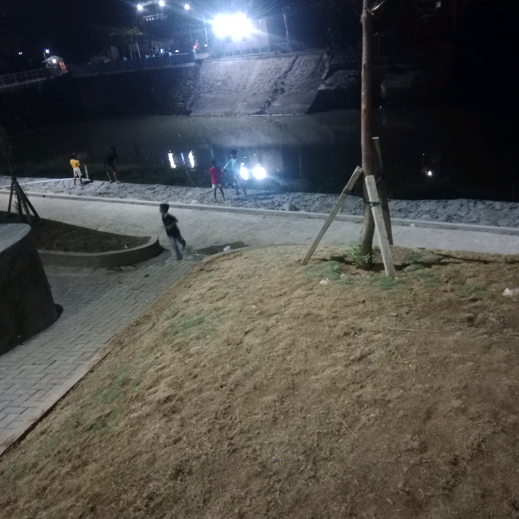 In Tlogorejo, Indonesia, at night, a scene unfolds near a body of water. The foreground is dominated by a sloping area covered in dry grass, with a paved walkway made of interlocking bricks bordering it on the left. Further ahead, a child runs along the walkway. Beyond the walkway, a group of three children stands by the water's edge. One child wears a yellow shirt and white shorts, another is in a red shirt, and the third is in a blue shirt. The water reflects the bright lights emanating from the background, where a wall of rocks forms the embankment. A wooden post with supporting braces stands near the center of the image, a solitary sentinel in the dim light.