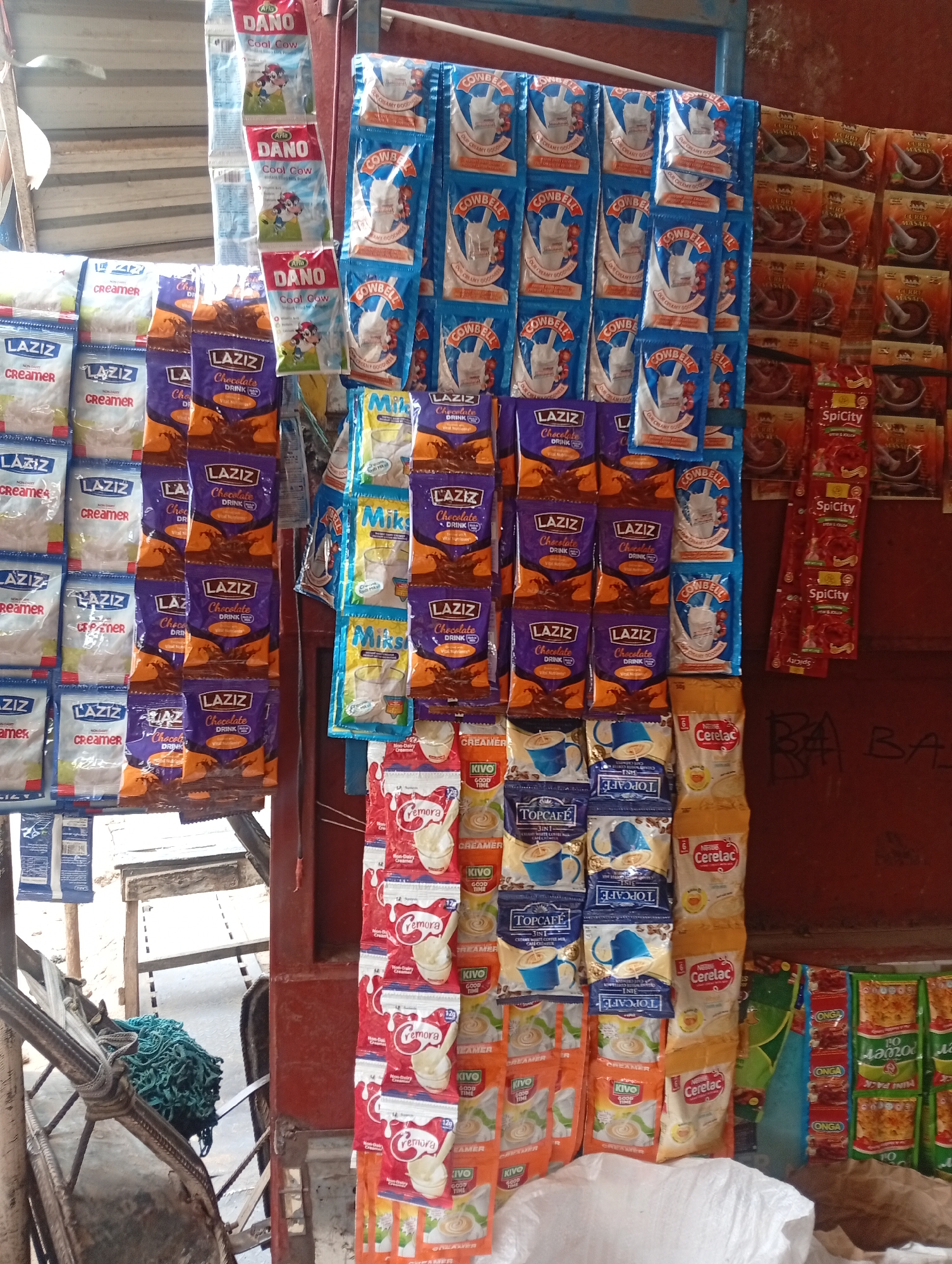 An outdoor retail kiosk in Sabon Tasha, Nigeria, displays an assortment of single-serving food and beverage packets. Ranging from milk powders like Cowbell and Dano to chocolate drinks from Laziz and Cerelac baby cereal, the products are hung in rows, primarily in blue, red, brown, white and yellow packaging. To the left, bags of Laziz creamer are on display. Behind the kiosk, a section of red corrugated metal is visible. The immediate foreground features part of a wheeled cart and a bag of white trash.