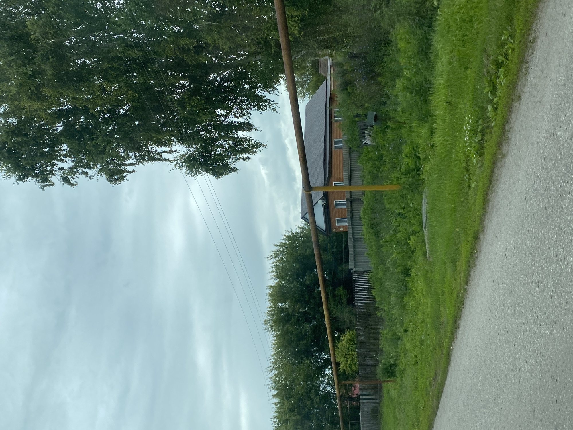 A rural scene in Nadezhdino, Russia, features a wooden house with a dark metal roof, bordered by a wooden fence. A large tree with green foliage stands to the right, with power lines visible in the overcast sky. A rusty metal pipe runs horizontally across the middle of the frame, supported by a yellow pole. The foreground shows a paved road and a grassy area with wildflowers. The overall impression is one of a quiet, lived-in rural landscape.