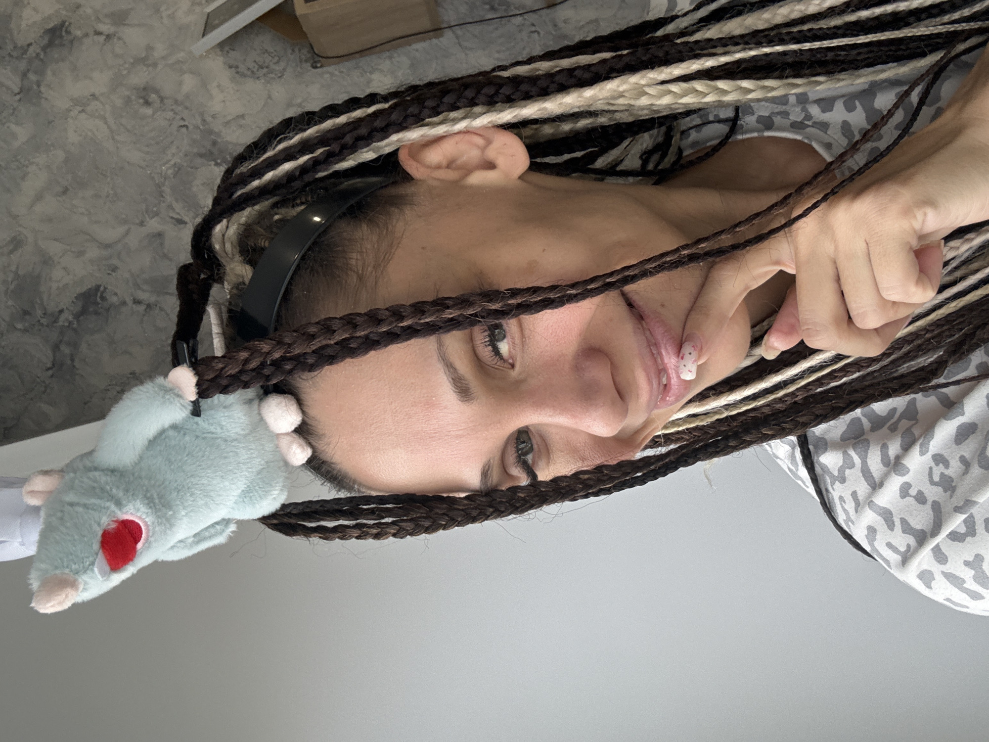 A rotated close-up depicts a light-skinned person with braided hair that includes dark brown, light brown, and white strands. The person is wearing a black headband and has a light-blue plush rat toy attached to it on the left side of the frame. The rat has a white snout, ears, and tail, with a red heart-shaped patch on its side. The person's facial features, including their eyes, nose, and mouth, are visible, and one hand is close to their mouth, with a nail design featuring white and red elements. The person is wearing a patterned shirt in shades of gray and white. The background is a solid gray, possibly a wall. The location is unknown.