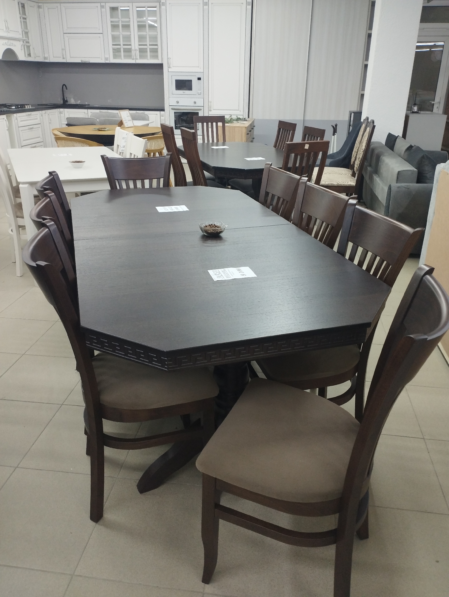 The image depicts a furniture showroom, likely located in Fălești, Moldova. The primary focus is on a dark wood dining table with a decorative edge, surrounded by several matching wooden chairs with upholstered seats. The table appears to be extended and set for a meal, with small white tags on its surface and a small glass dish containing a brown substance in the center. In the background, more furniture is visible, including kitchen cabinets, a sofa, and other tables and chairs, suggesting a retail environment. The flooring is tiled in a neutral color. There are no people visible in the image.