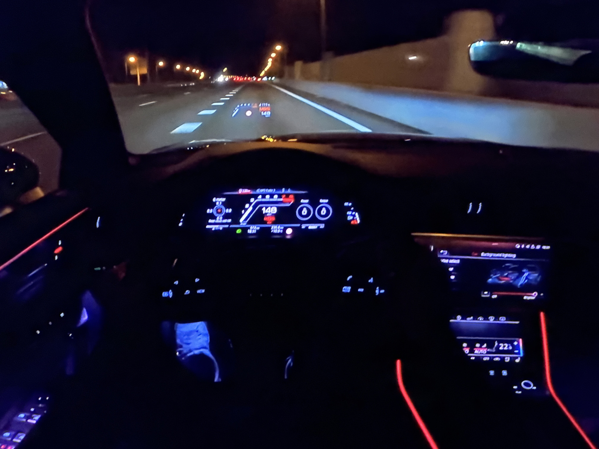 The image captures a nighttime view from the driver's seat of a car on a highway, possibly near Bezden, Bulgaria. The dashboard displays illuminated digital readouts, including a speed of 148, and a head-up display (HUD) projects speed and navigation information onto the windshield. Red and blue ambient lighting accents the interior. The road ahead is illuminated by streetlights and the car's headlights, revealing lane markings and the edge of the road. The car appears to be in motion.